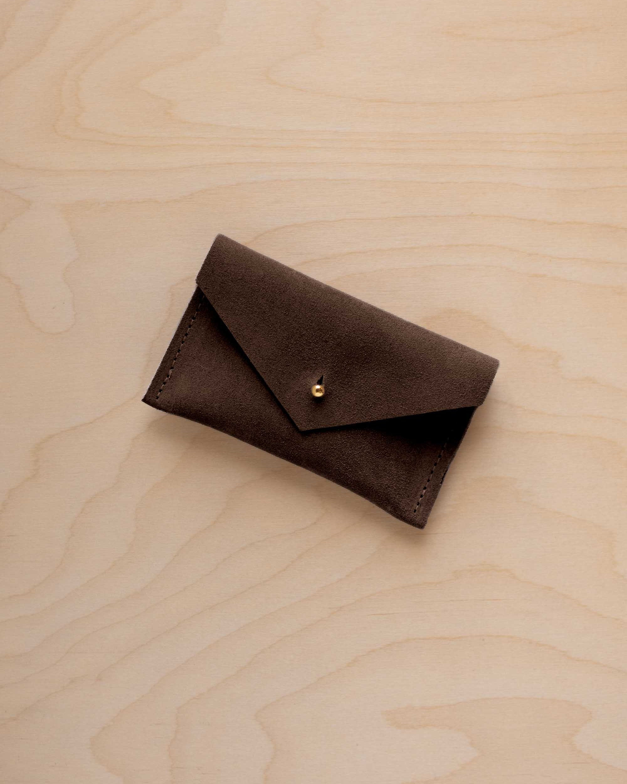 A small espresso brown coloured suede pouch with a flap closure and gold coloured fastening, on a light wood grain surface.