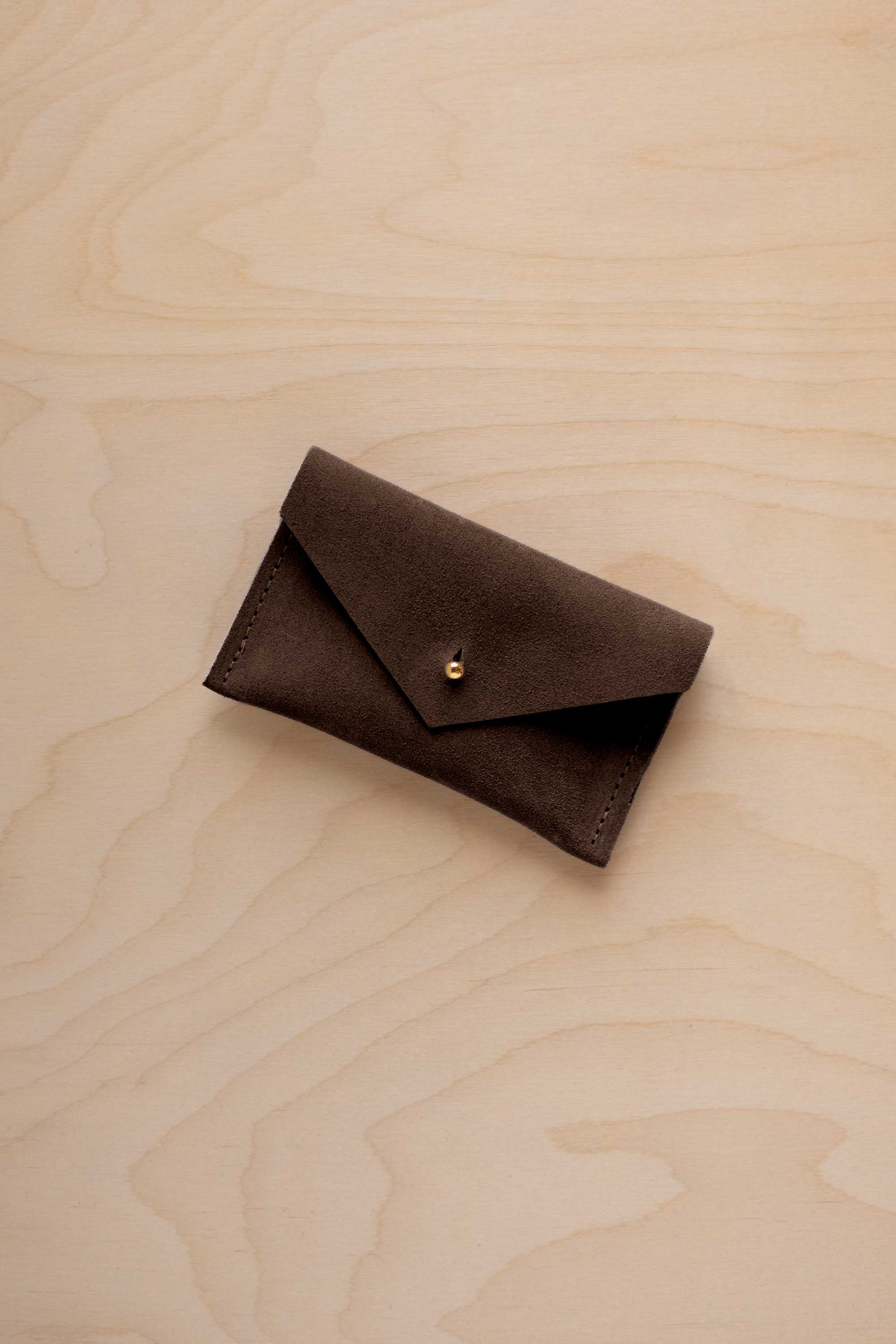 A small espresso brown coloured suede pouch with a flap closure and gold coloured fastening, on a light wood grain surface.
