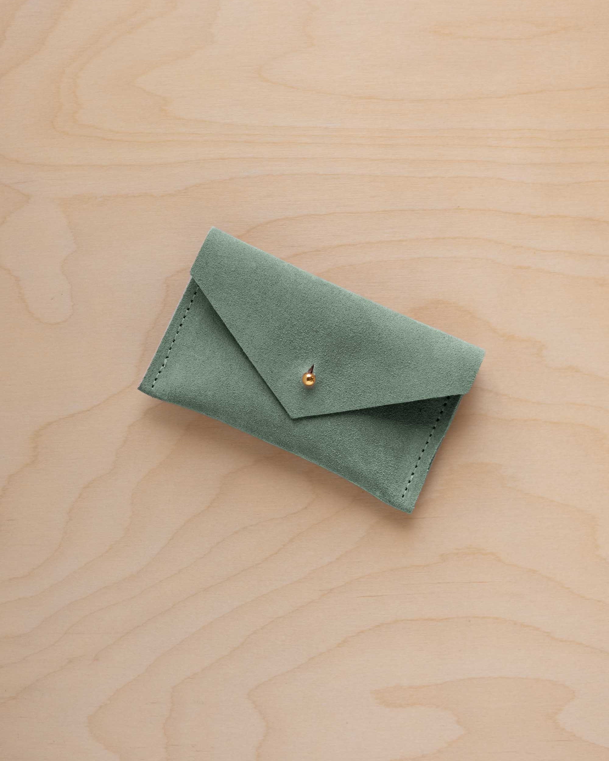 A small eucalyptus green coloured suede pouch with a flap closure and gold coloured fastening, on a light wood grain surface.