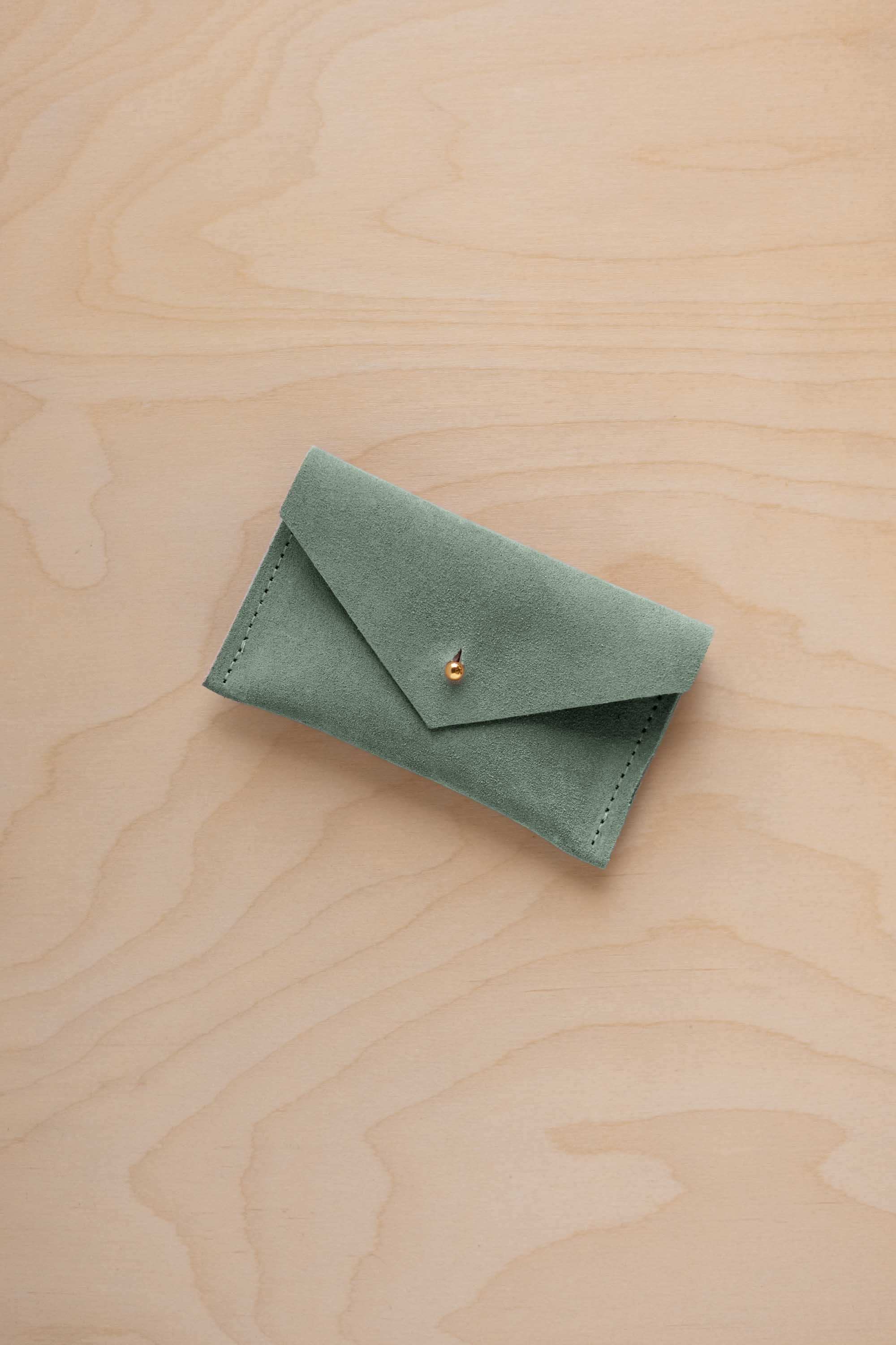 A small eucalyptus green coloured suede pouch with a flap closure and gold coloured fastening, on a light wood grain surface.