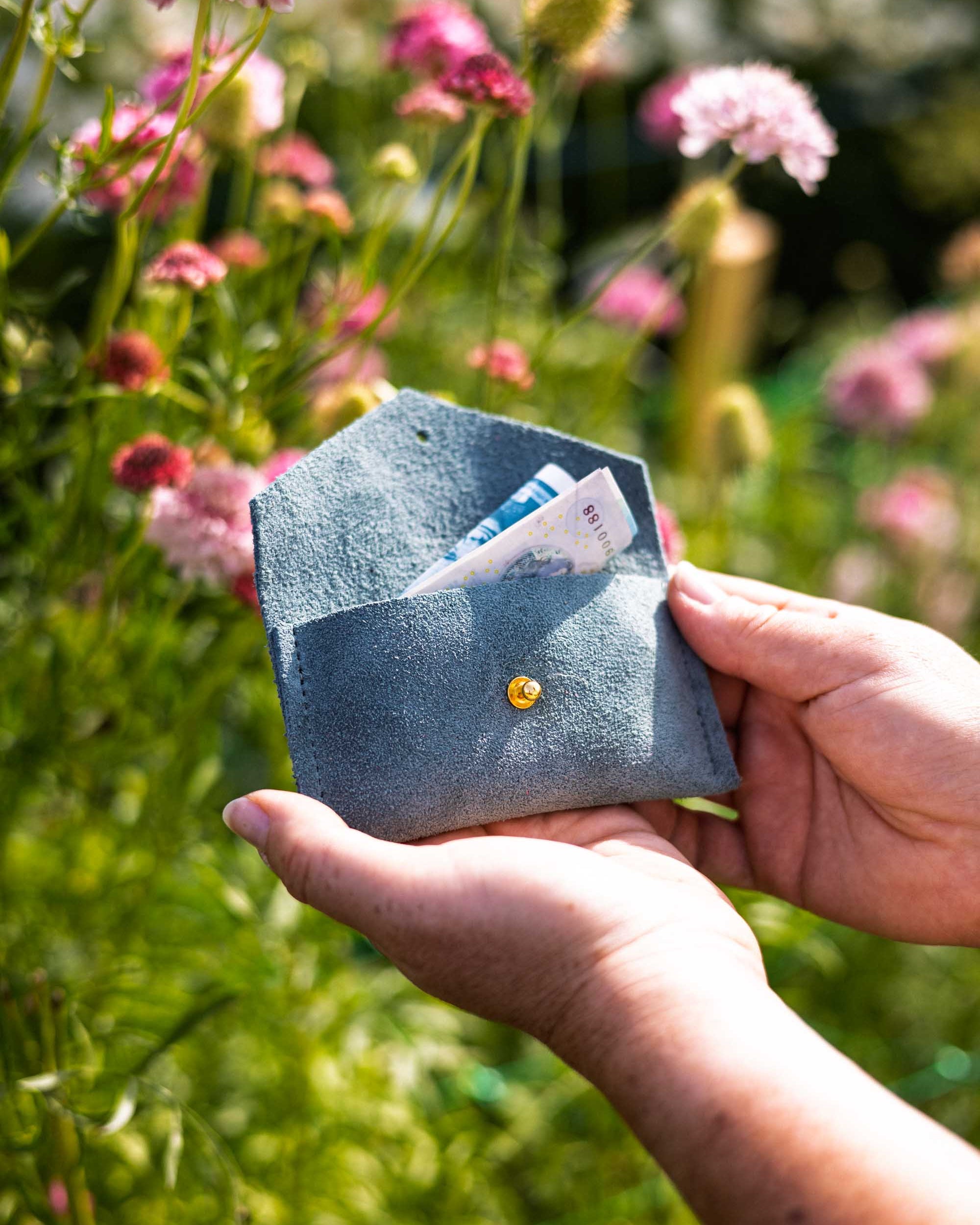 A person's hands holding an open eucalyptus or sage green envelope-style wallet with a gold-tone button, showing a folded money notes inside, set against a blurred background of green foliage and pink wildflowers.