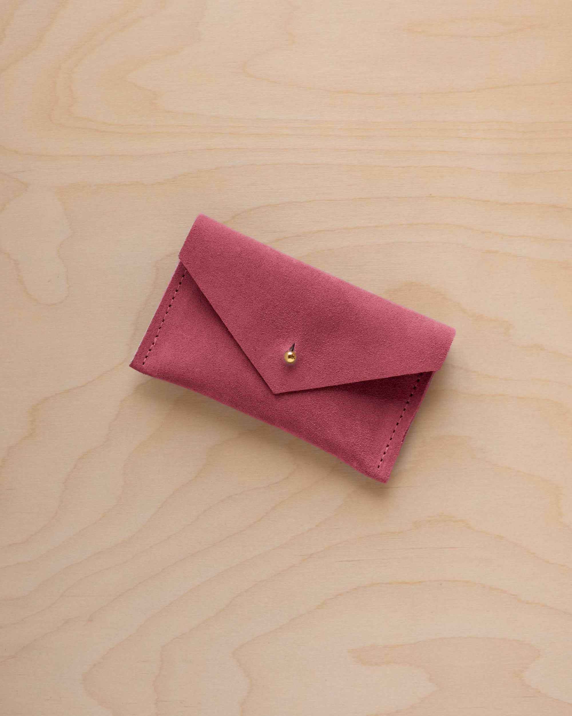 A small faded rose coloured suede pouch with a flap closure and gold coloured fastening, on a light wood grain surface.