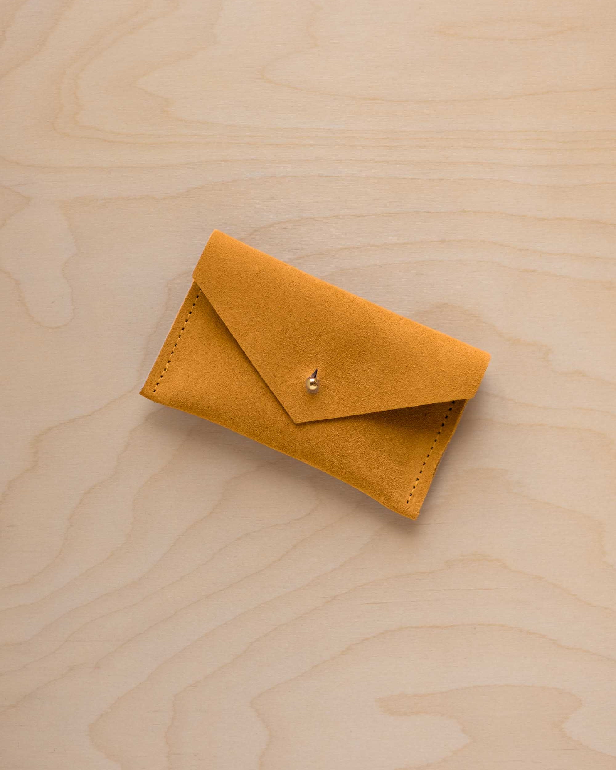 A small golden ochre coloured suede pouch with a flap closure and gold coloured fastening, on a light wood grain surface.