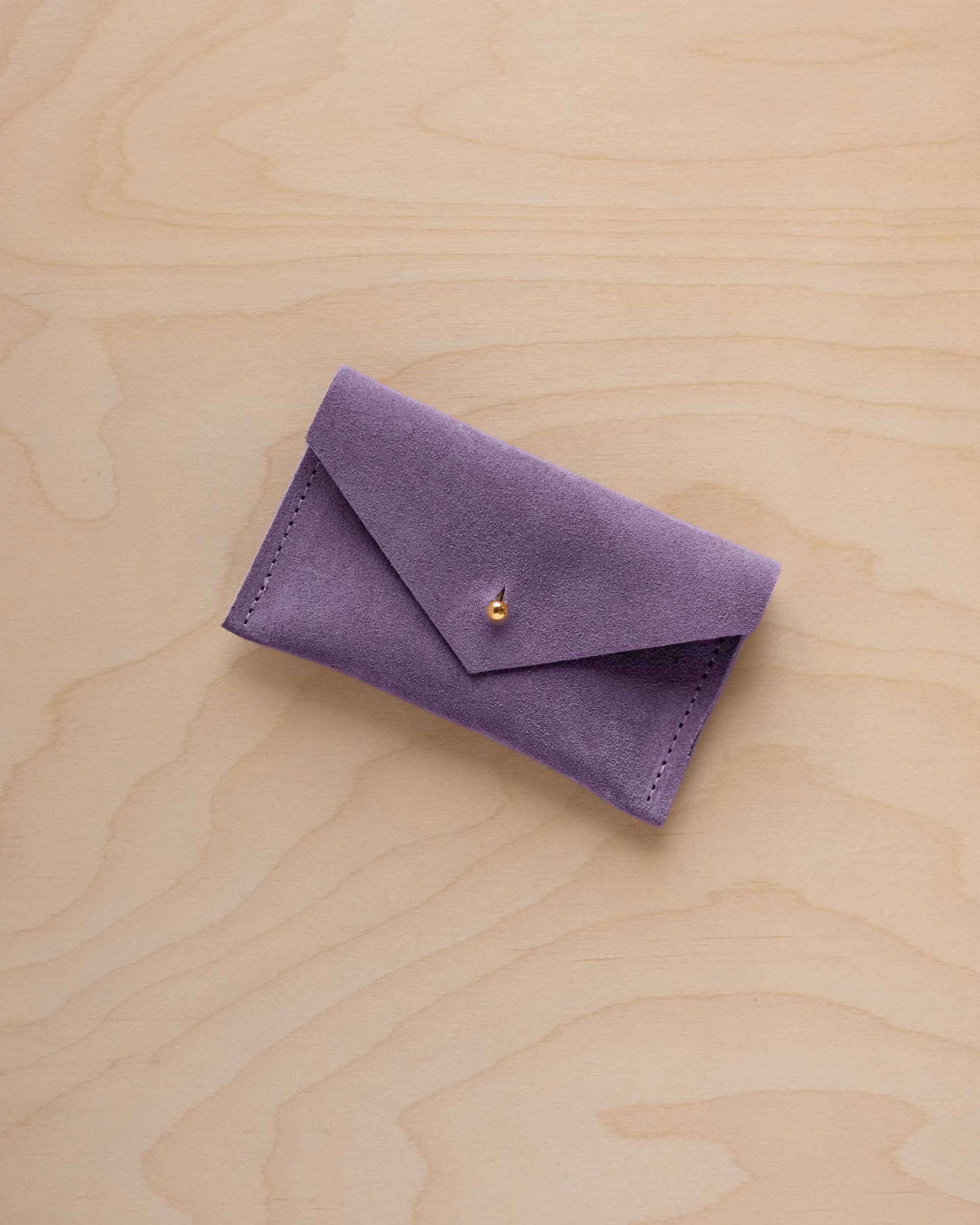 A small lavender coloured suede pouch with a flap closure and gold coloured fastening, on a light wood grain surface.