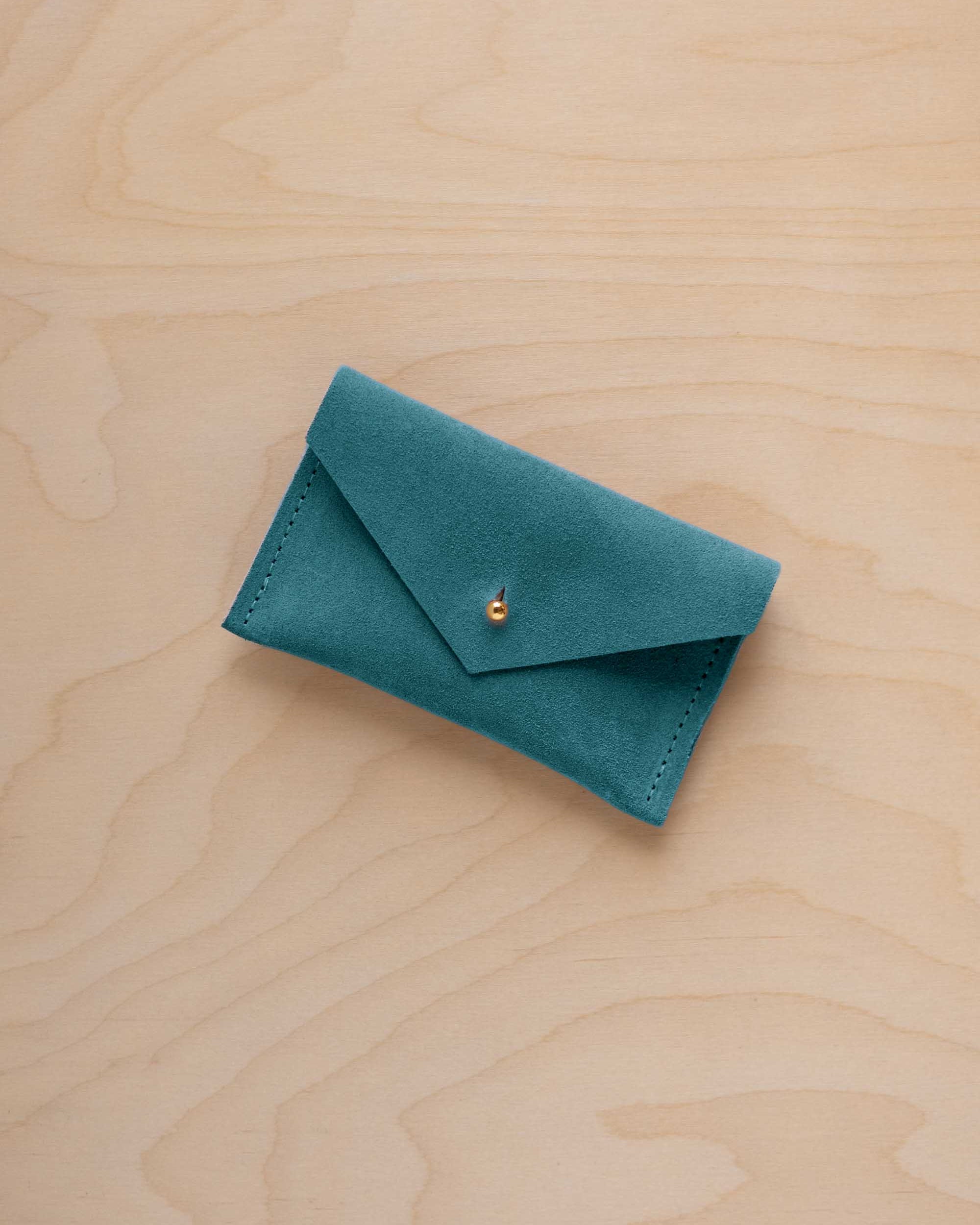 A small marine teal coloured suede pouch with a flap closure and gold coloured fastening, on a light wood grain surface.