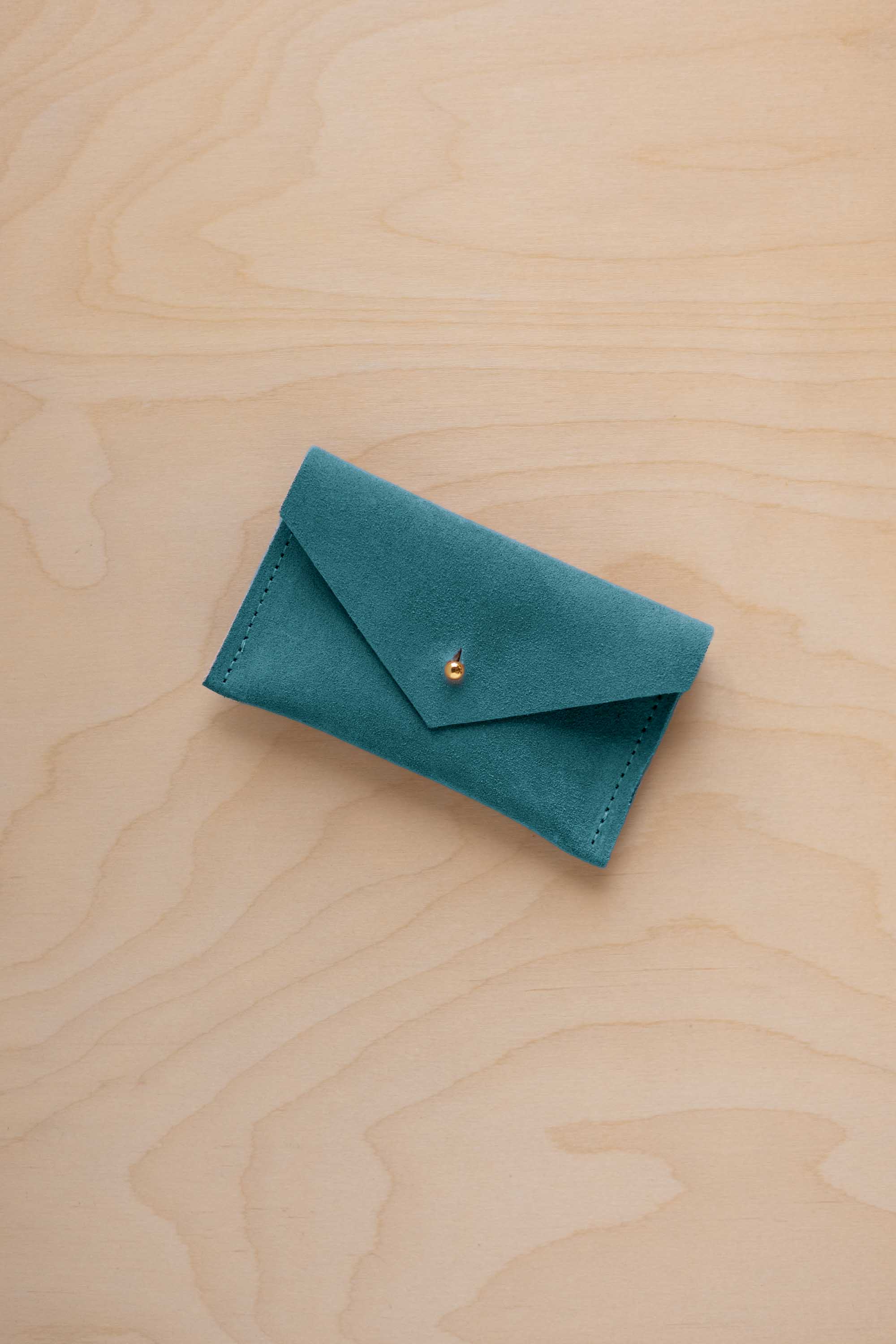 A small marine teal coloured suede pouch with a flap closure and gold coloured fastening, on a light wood grain surface.