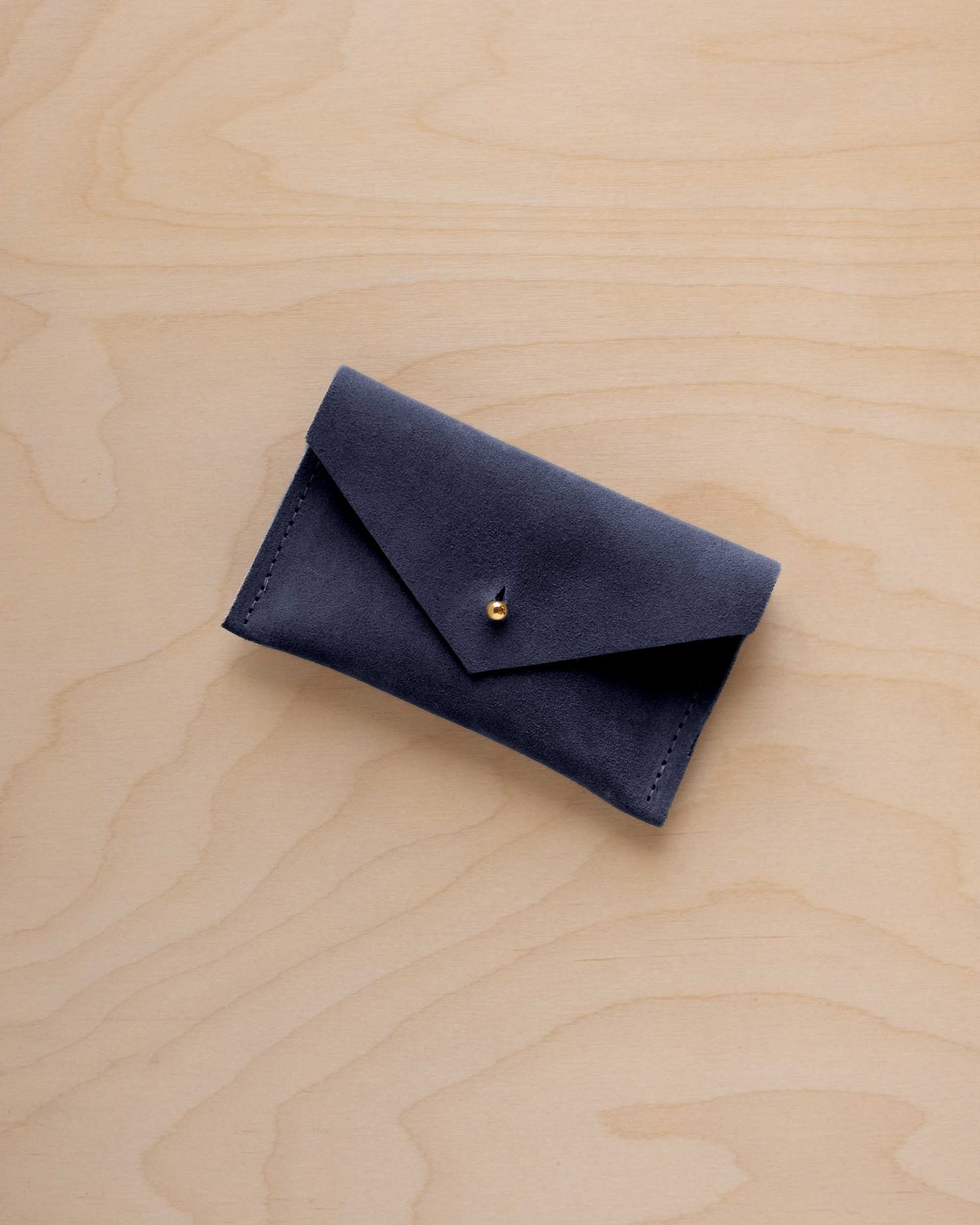A small midnight navy coloured suede pouch with a flap closure and gold coloured fastening, on a light wood grain surface.