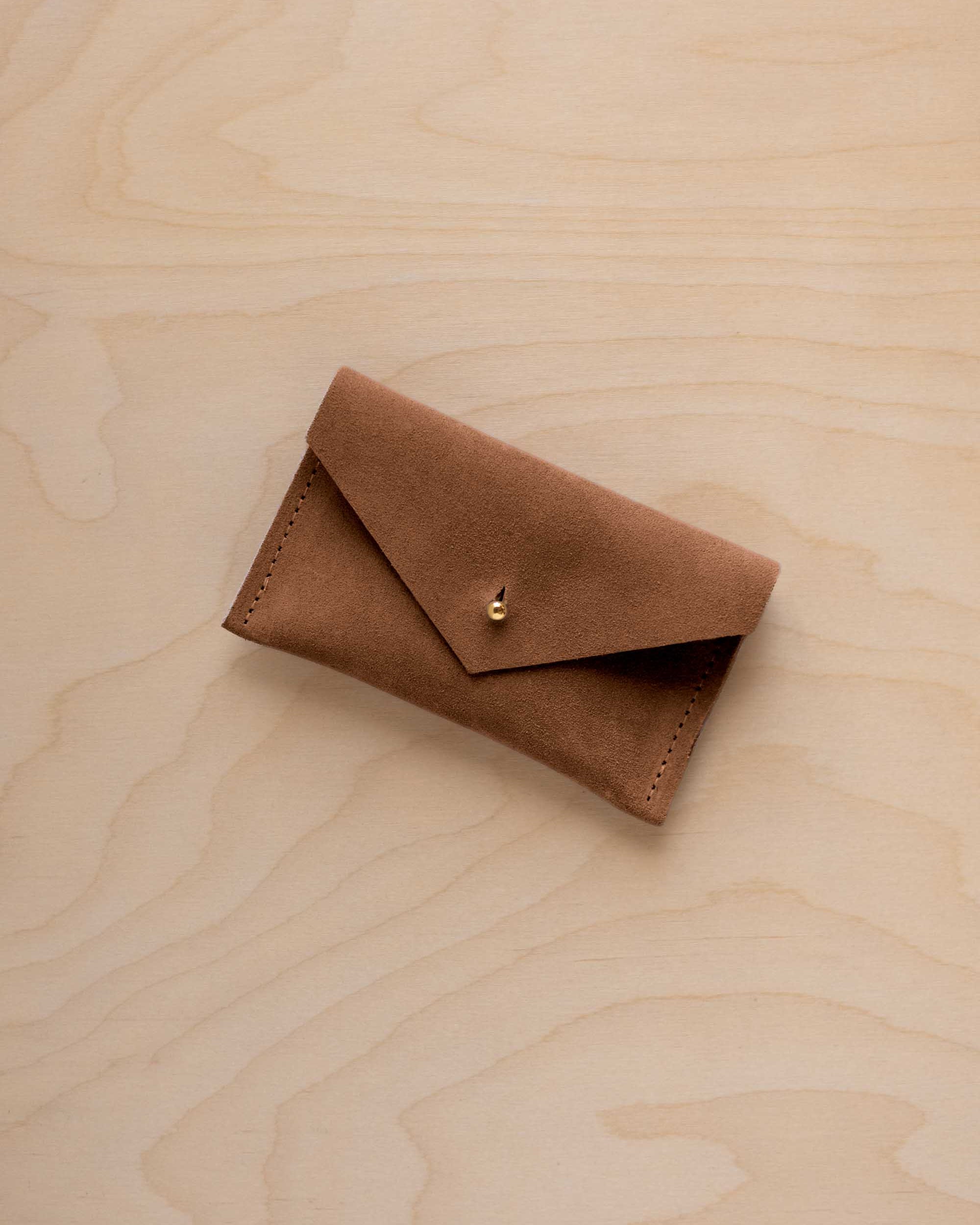 A small mocha brown coloured suede pouch with a flap closure and gold coloured fastening, on a light wood grain surface.