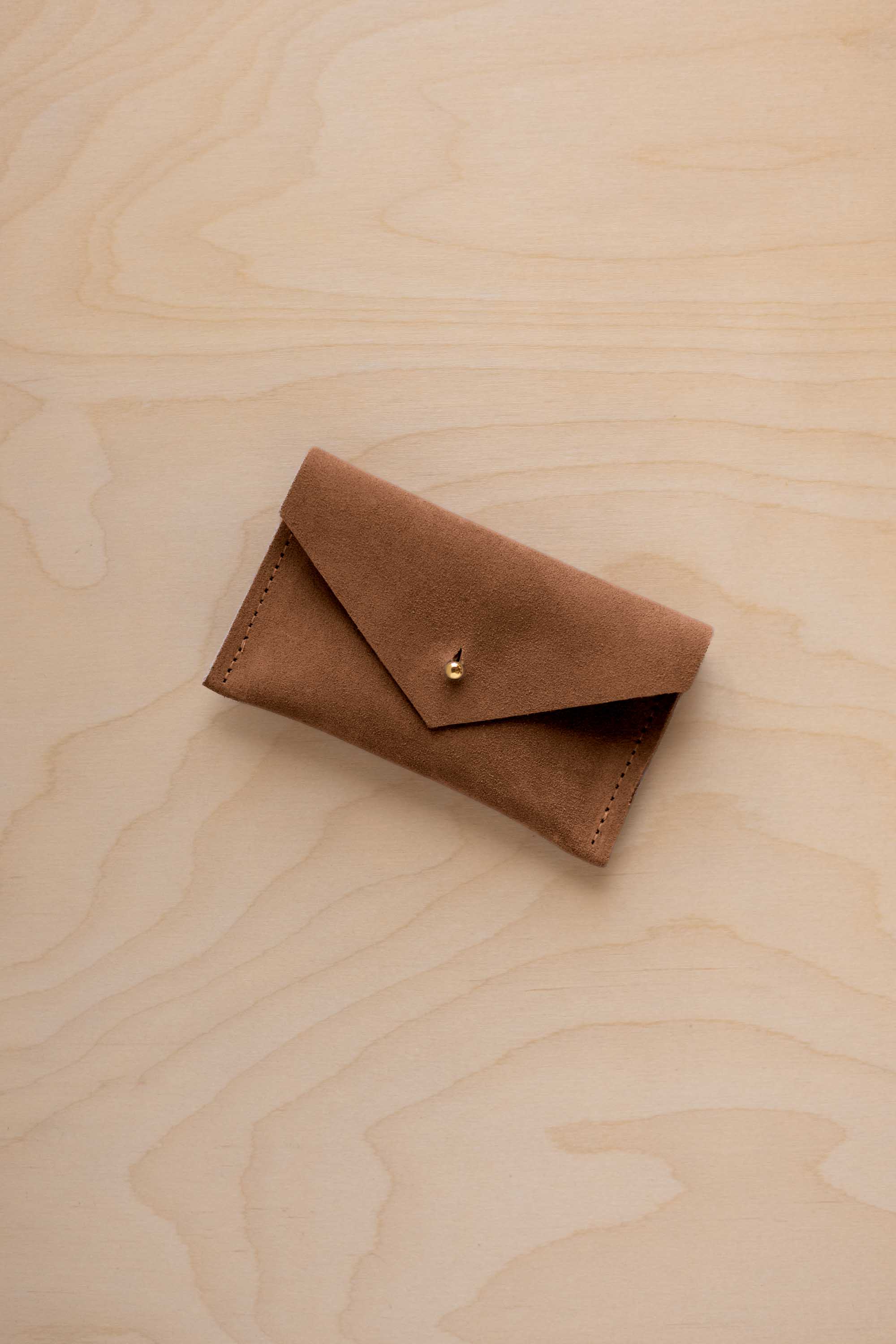 A small mocha brown coloured suede pouch with a flap closure and gold coloured fastening, on a light wood grain surface.