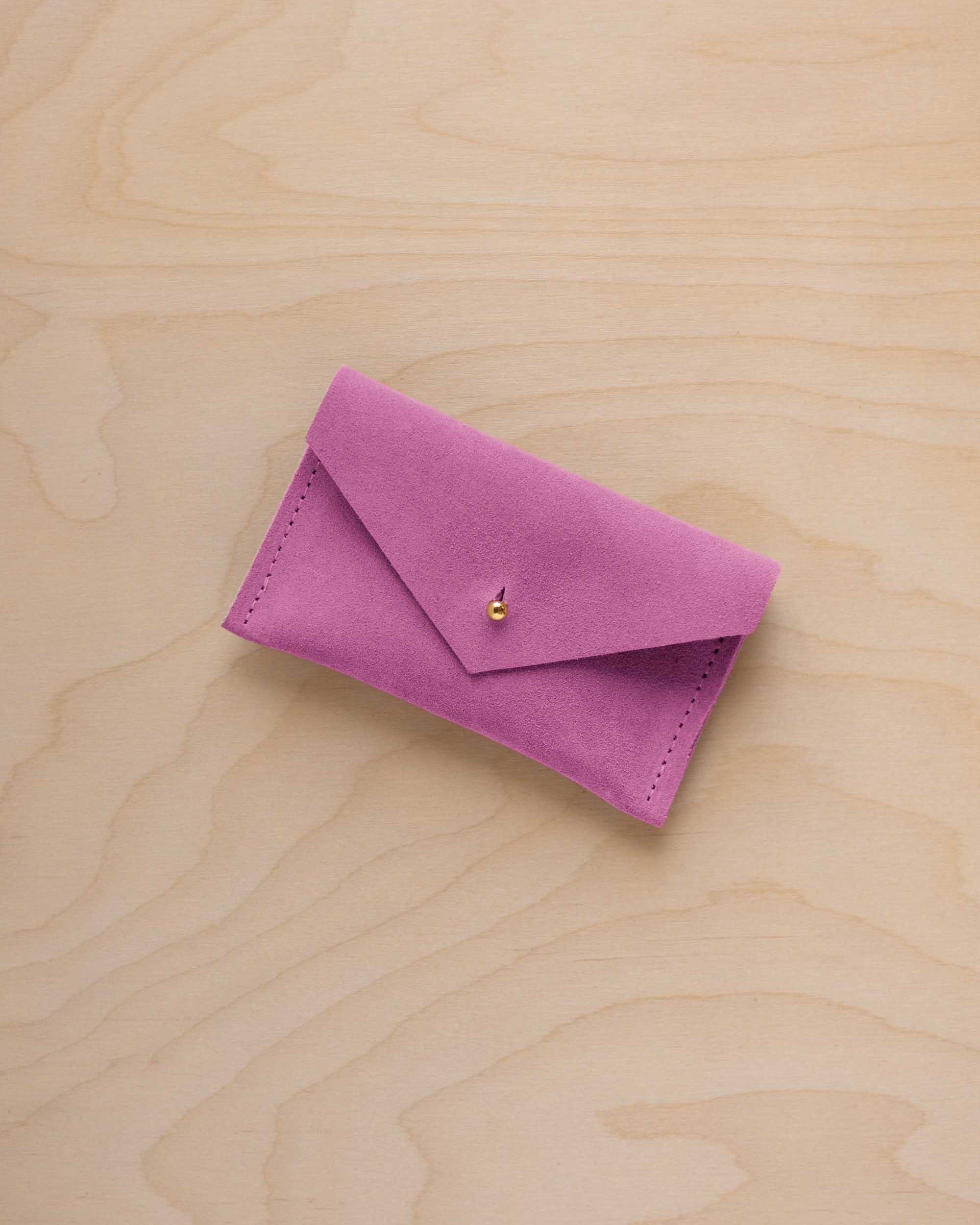 A small peony pink coloured suede pouch with a flap closure and gold coloured fastening, on a light wood grain surface.