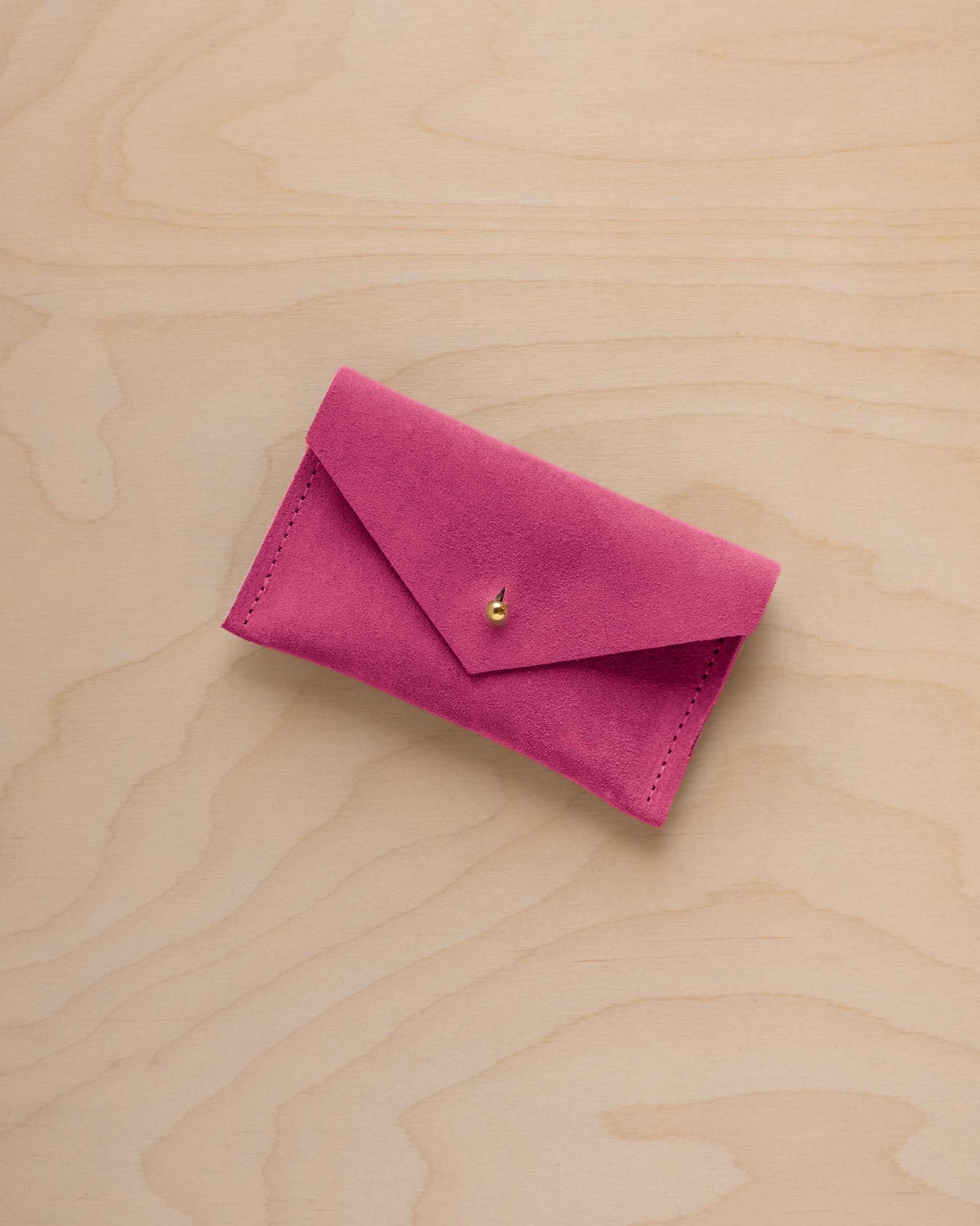A small raspberry coloured suede pouch with a flap closure and gold coloured fastening, on a light wood grain surface.