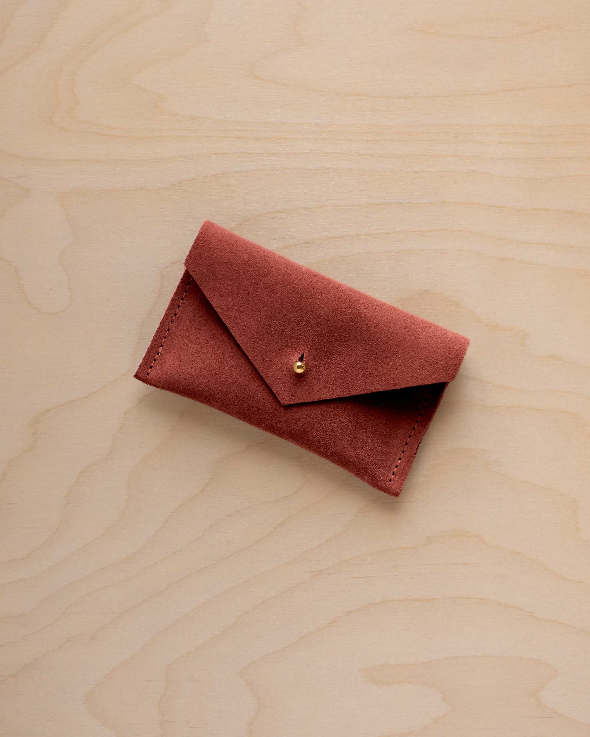 A small rust red coloured suede pouch with a flap closure and gold coloured fastening, on a light wood grain surface.