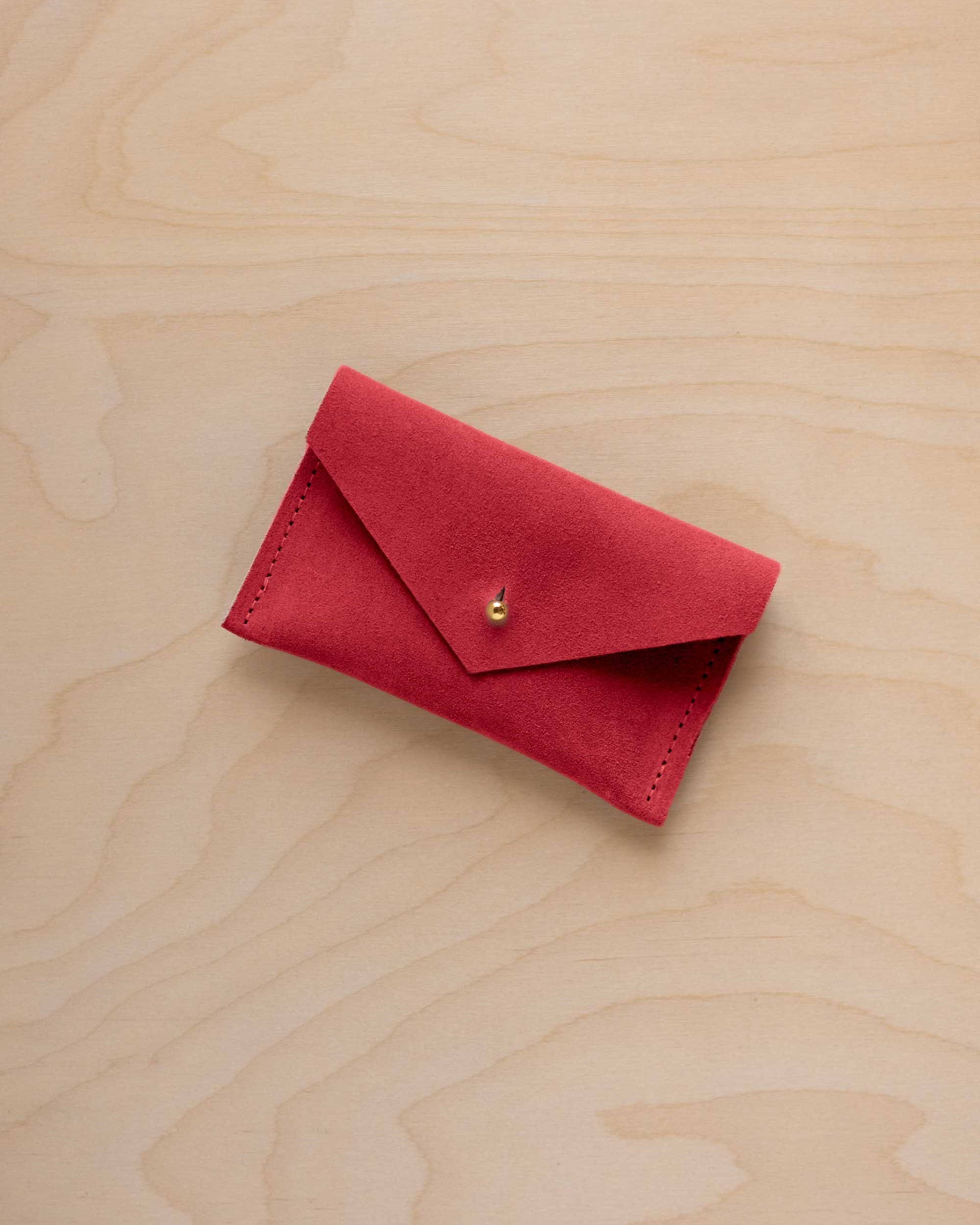 A small scarlet red coloured suede pouch with a flap closure and gold coloured fastening, on a light wood grain surface.