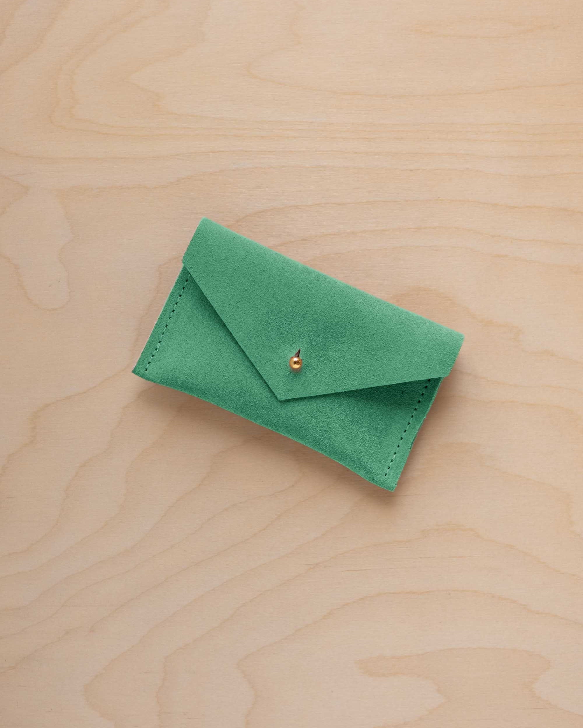 A small seafoam green coloured suede pouch with a flap closure and gold coloured fastening, on a light wood grain surface.