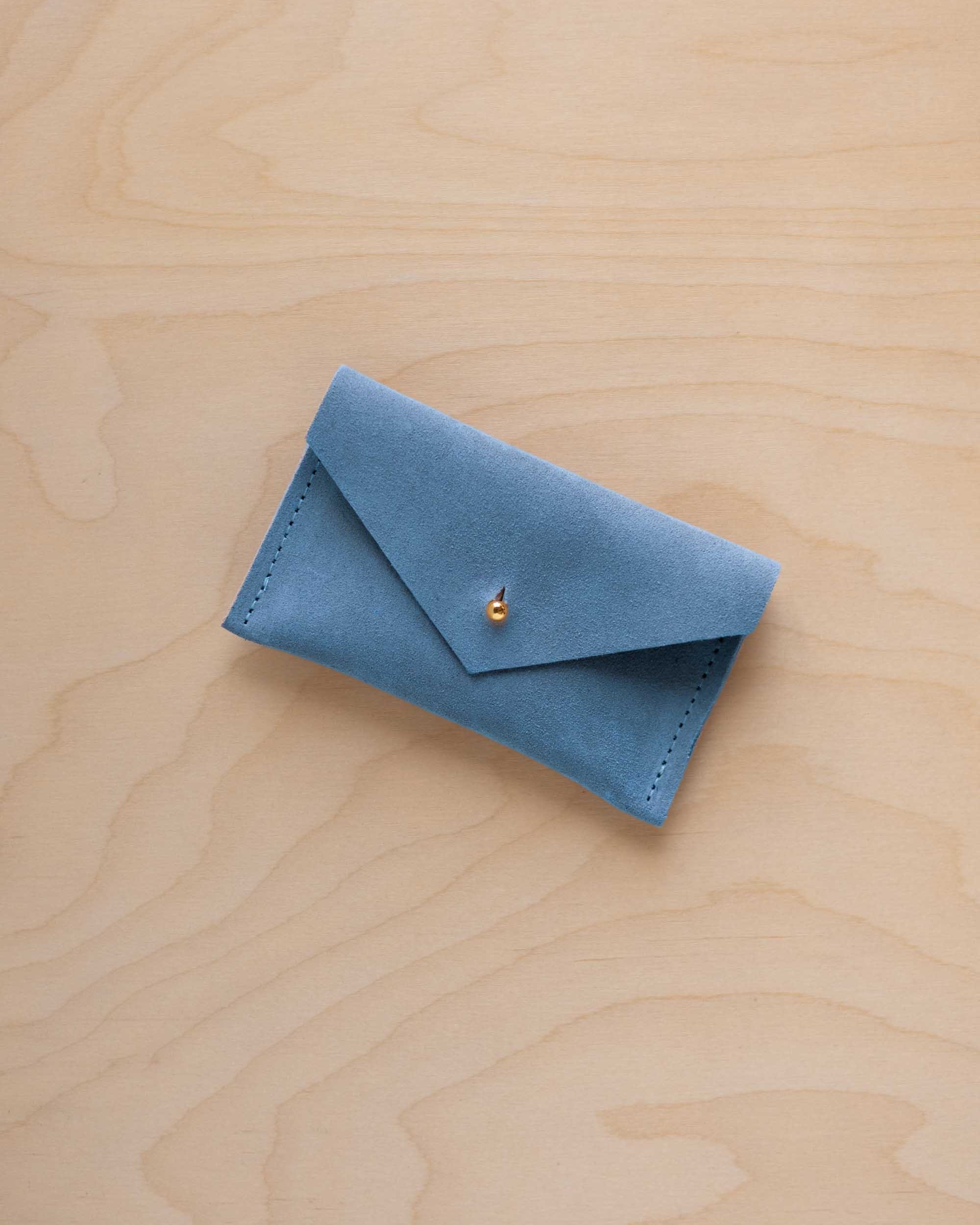 A small sky blue coloured suede pouch with a flap closure and gold coloured fastening, on a light wood grain surface.