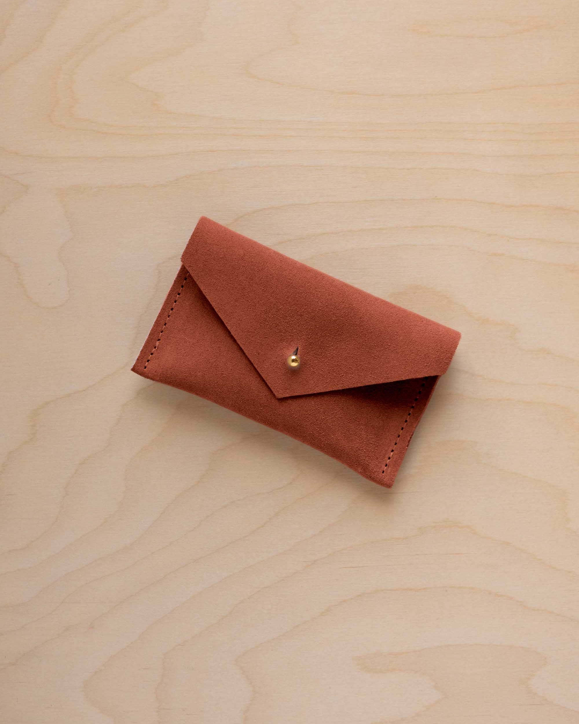 A small terracotta coloured suede pouch with a flap closure and gold coloured fastening, on a light wood grain surface.