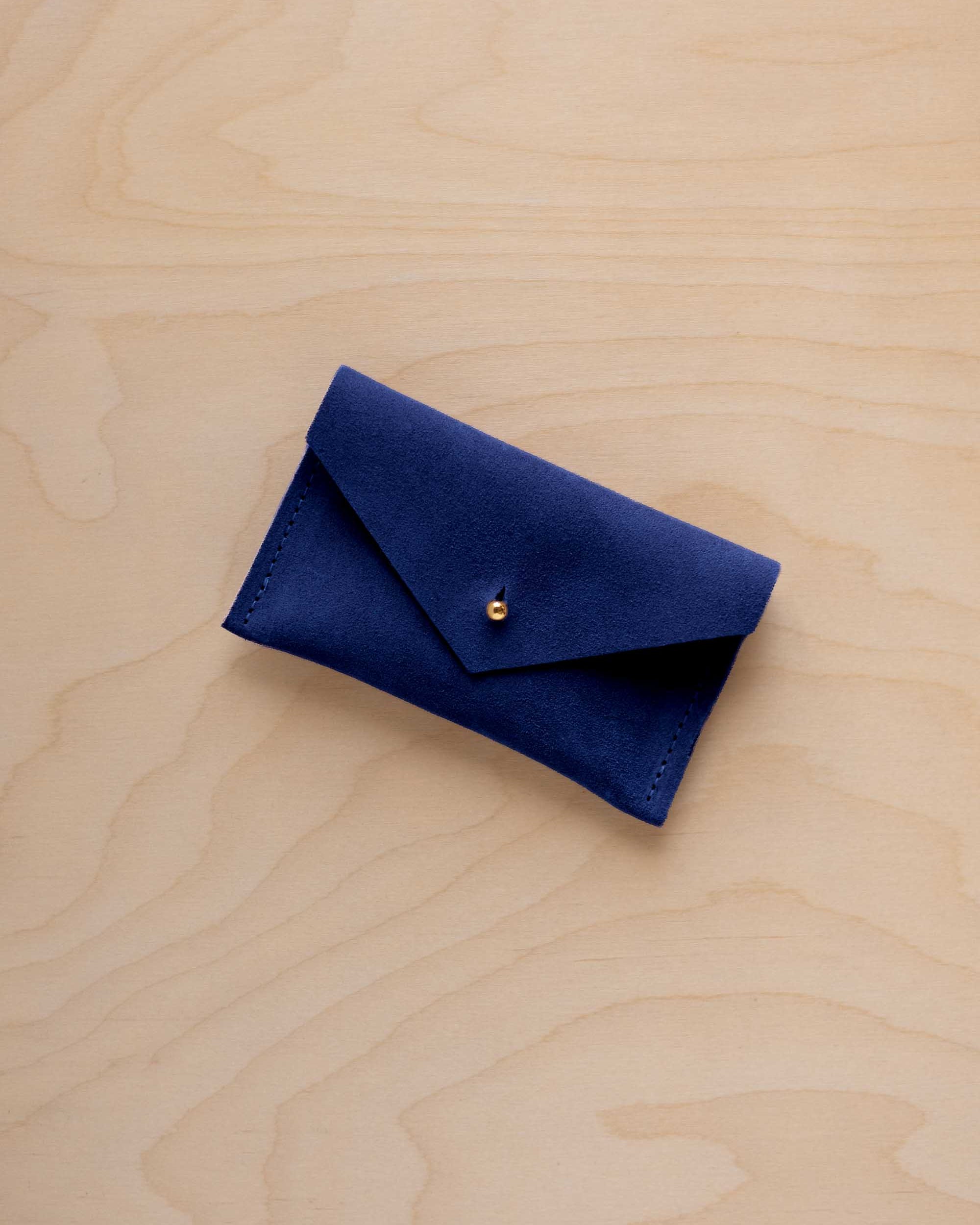 A small ultramarine blue coloured suede pouch with a flap closure and gold coloured fastening, on a light wood grain surface.
