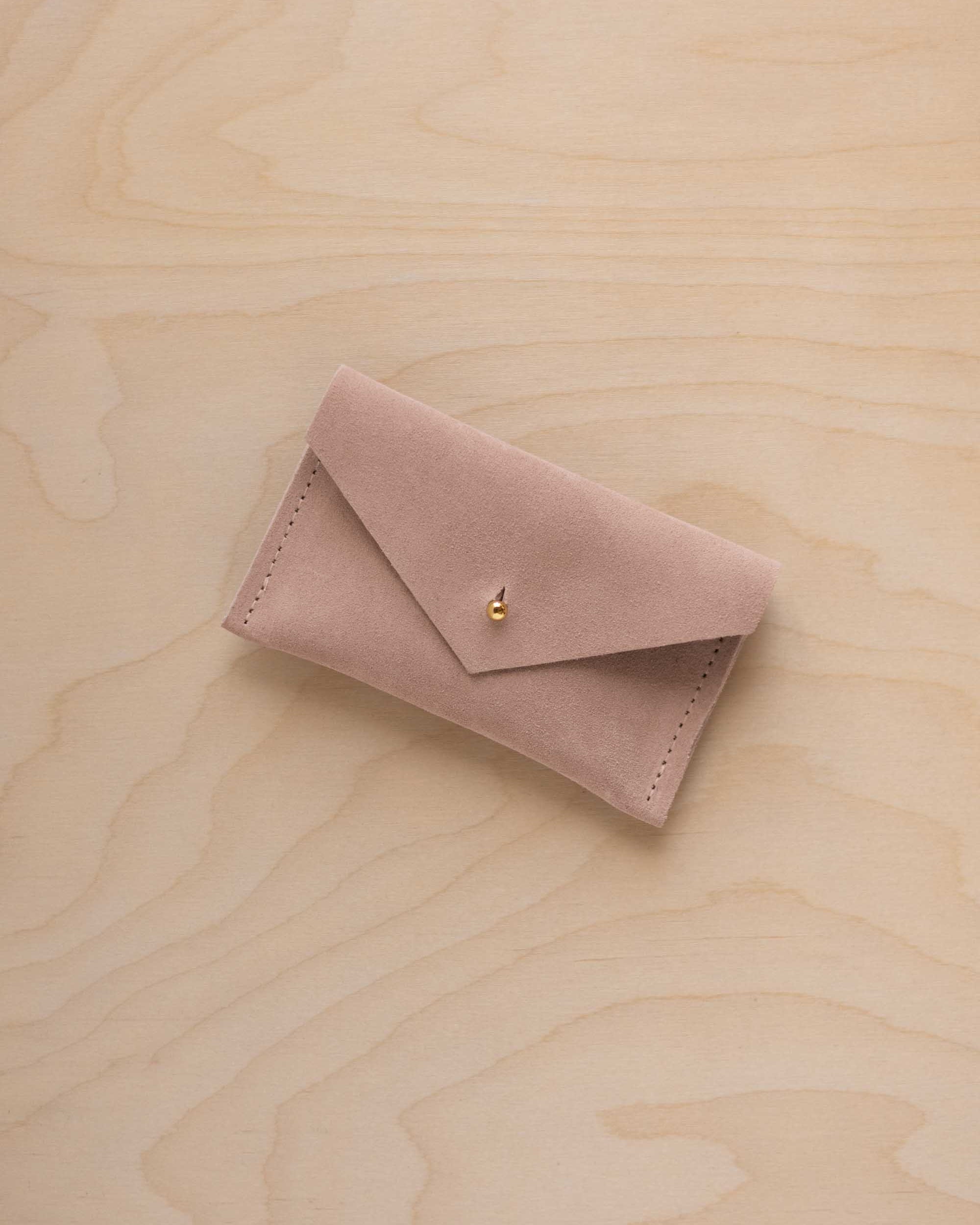 A small warm sand coloured suede pouch with a flap closure and gold button, on a light wood grain surface.