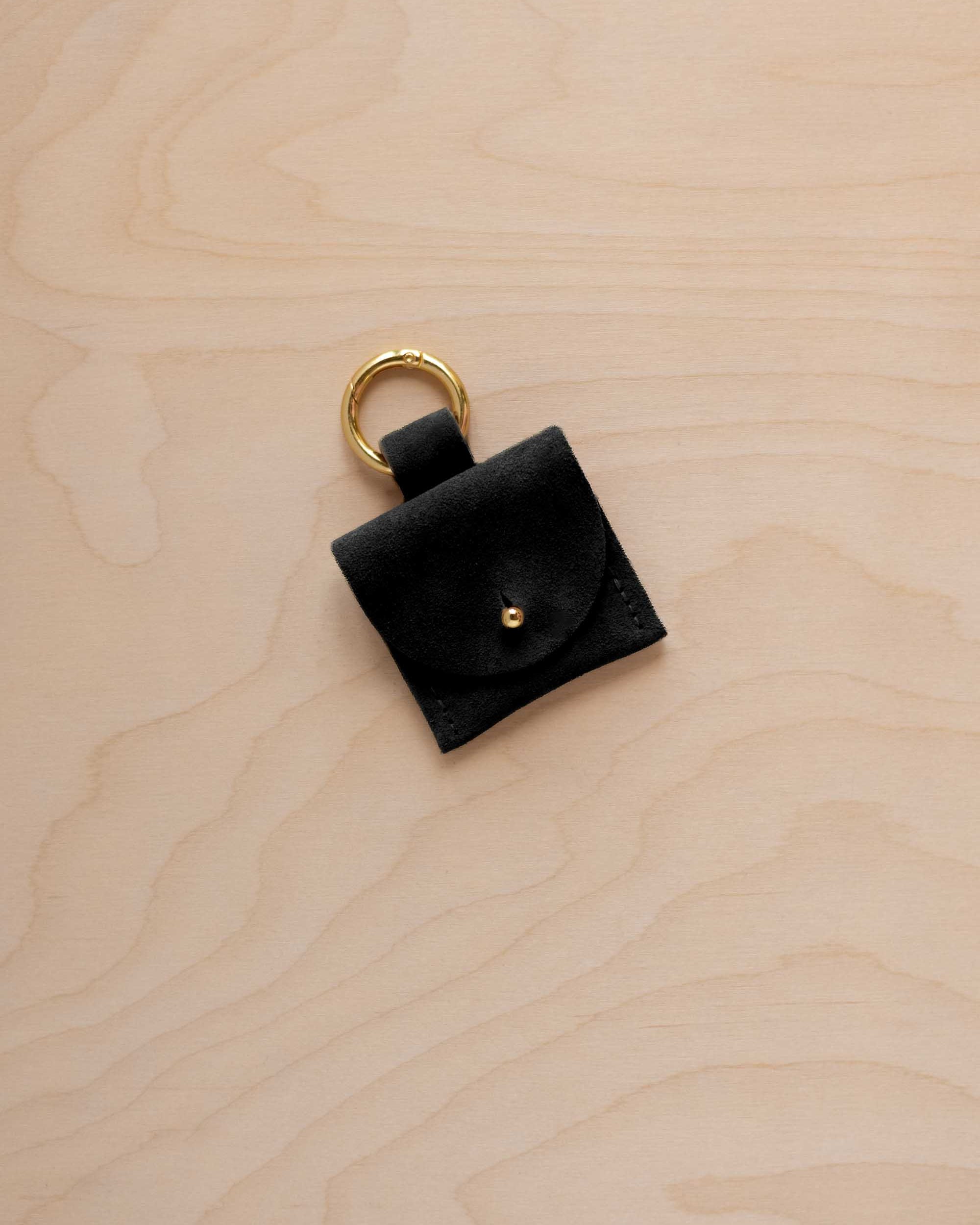 A small black suede pouch bag charm with a gold coloured clip ring fastening, displayed on a light wood grain background.