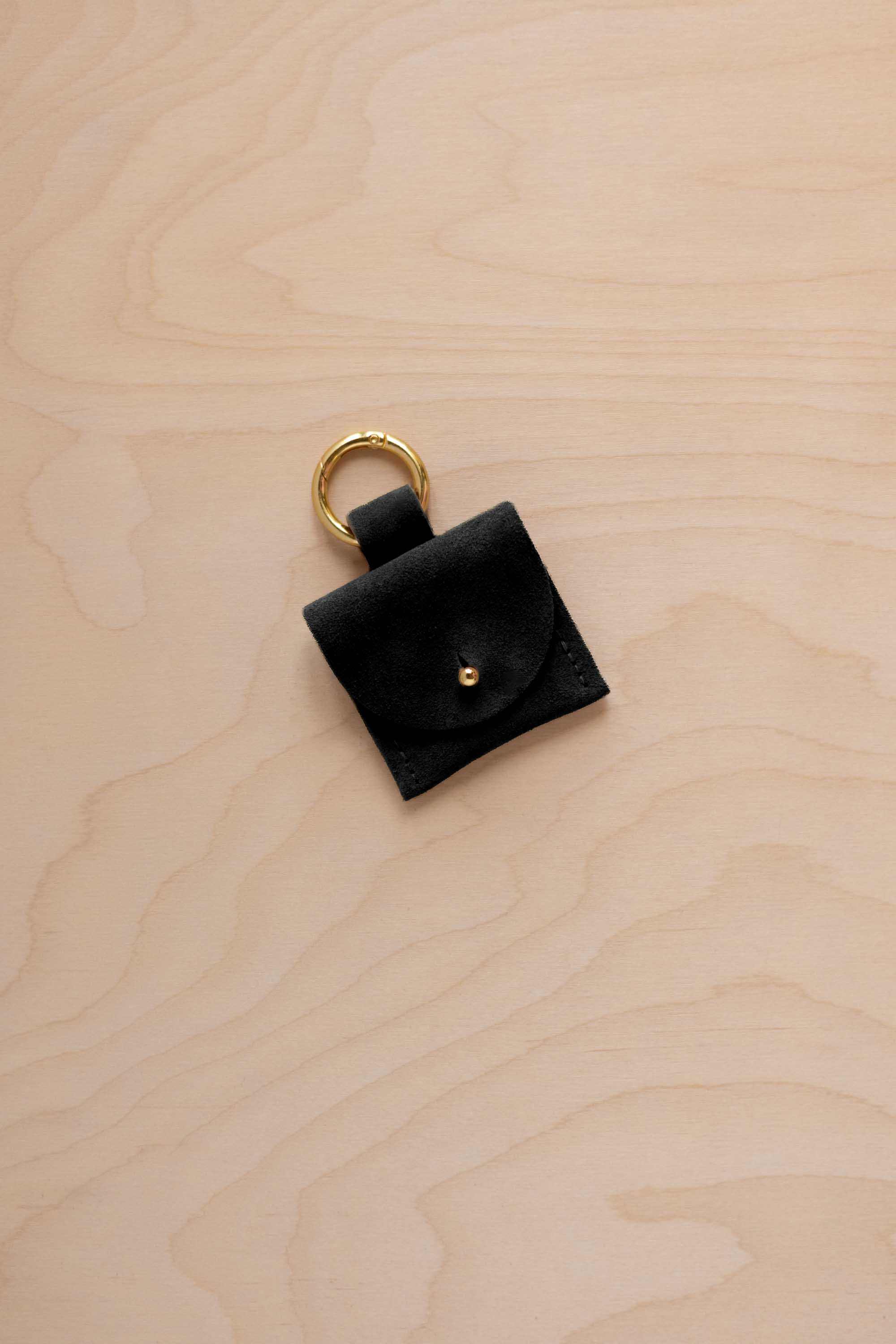 A small black suede pouch bag charm with a gold coloured clip ring fastening, displayed on a light wood grain background.