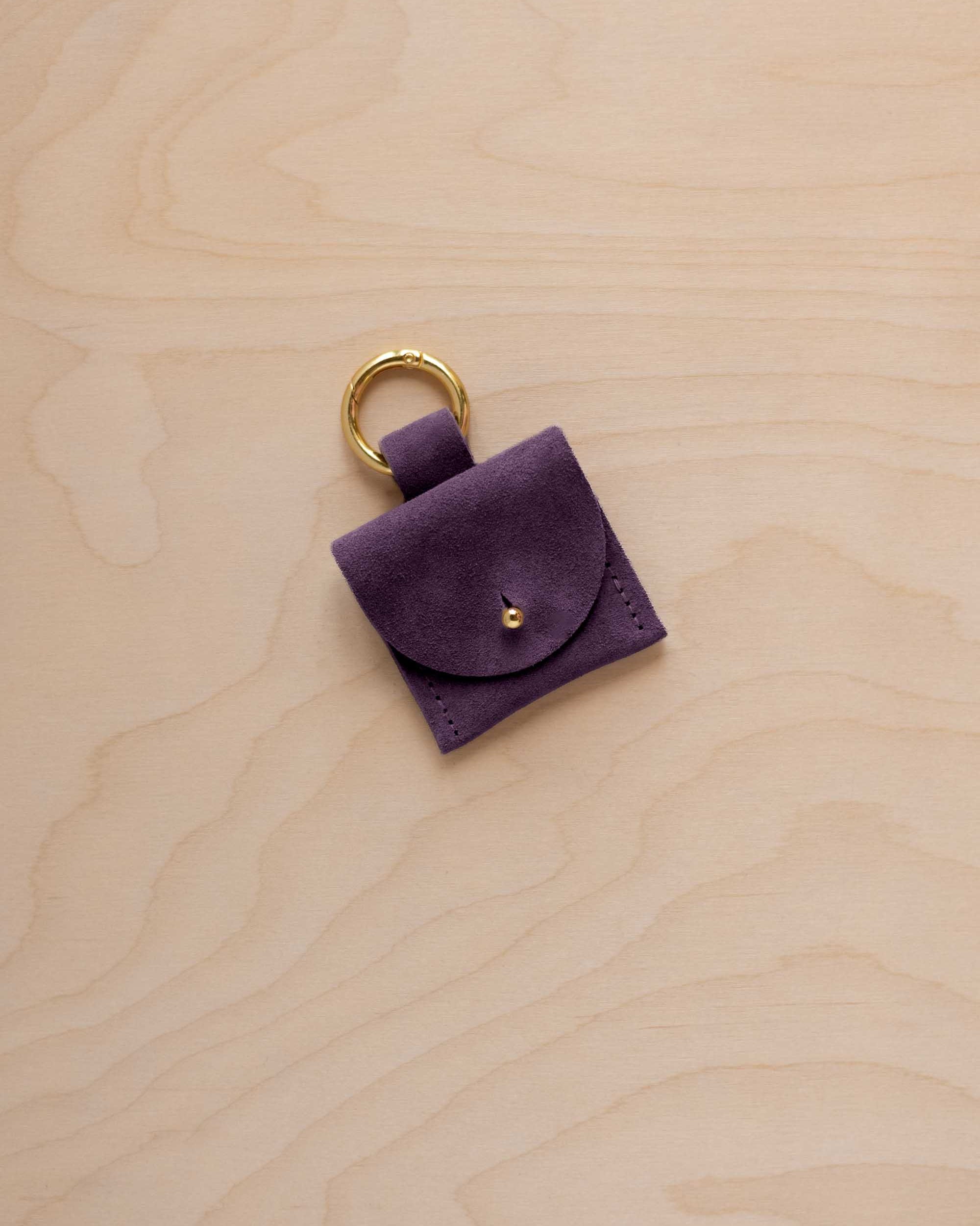 A small blackcurrant dark purple suede pouch bag charm with a gold coloured clip ring fastening, displayed on a light wood grain background.