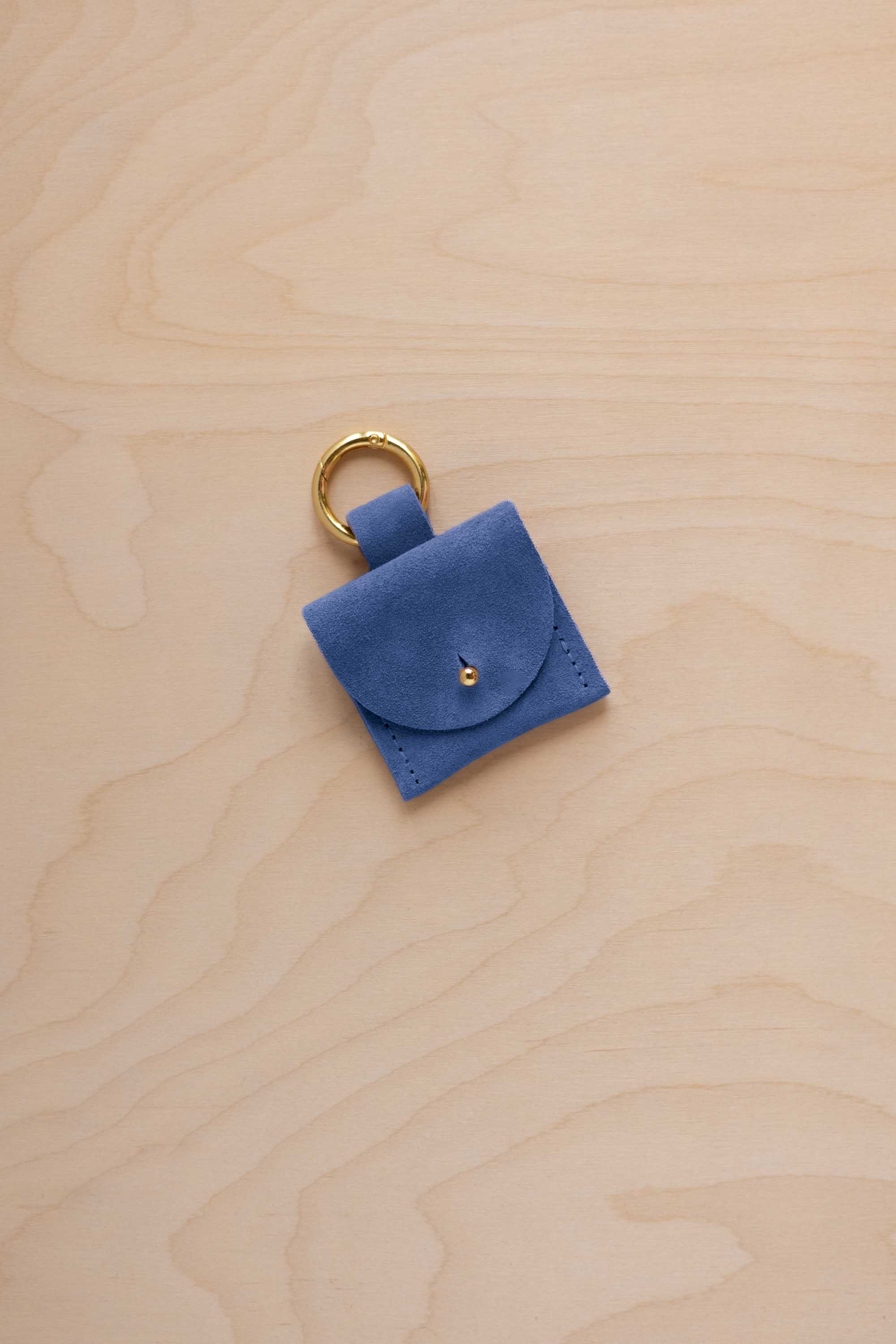 A small cobalt blue coloured suede pouch bag charm with a gold coloured clip ring fastening, displayed on a light wood grain background.