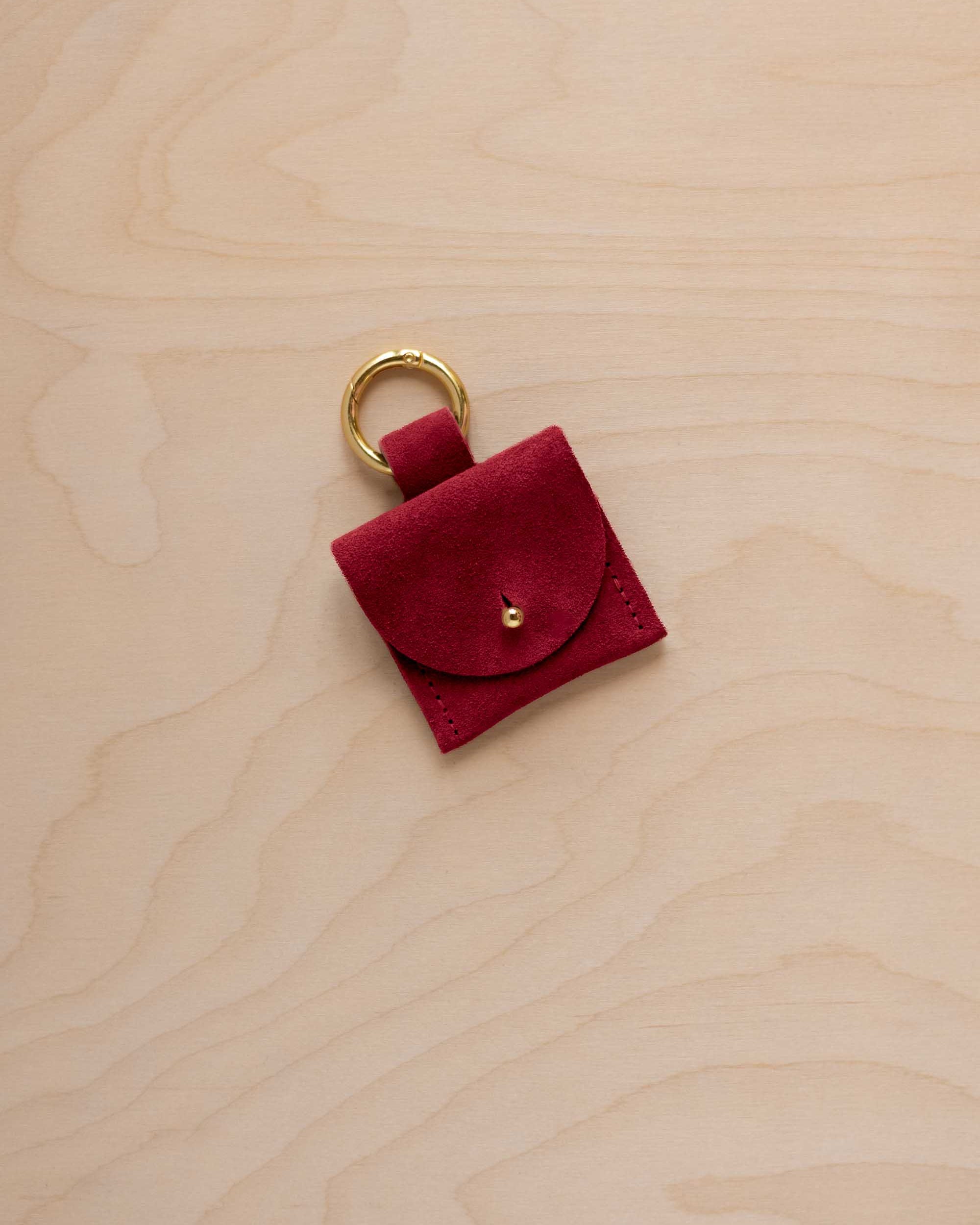 A small crimson red coloured suede pouch bag charm with a gold coloured clip ring fastening, displayed on a light wood grain background.