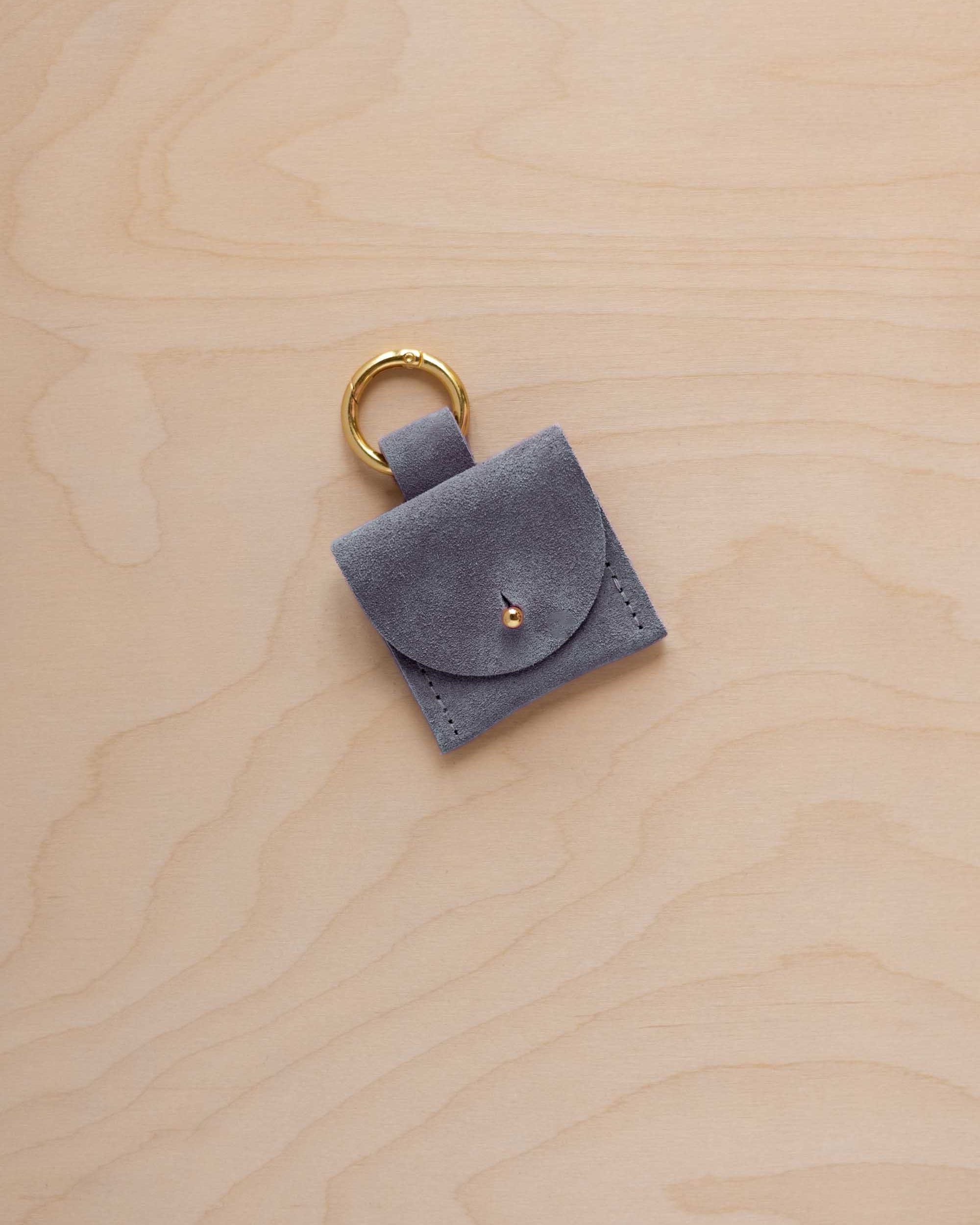 A small dusky blue coloured suede pouch bag charm with a gold coloured clip ring fastening, displayed on a light wood grain background.