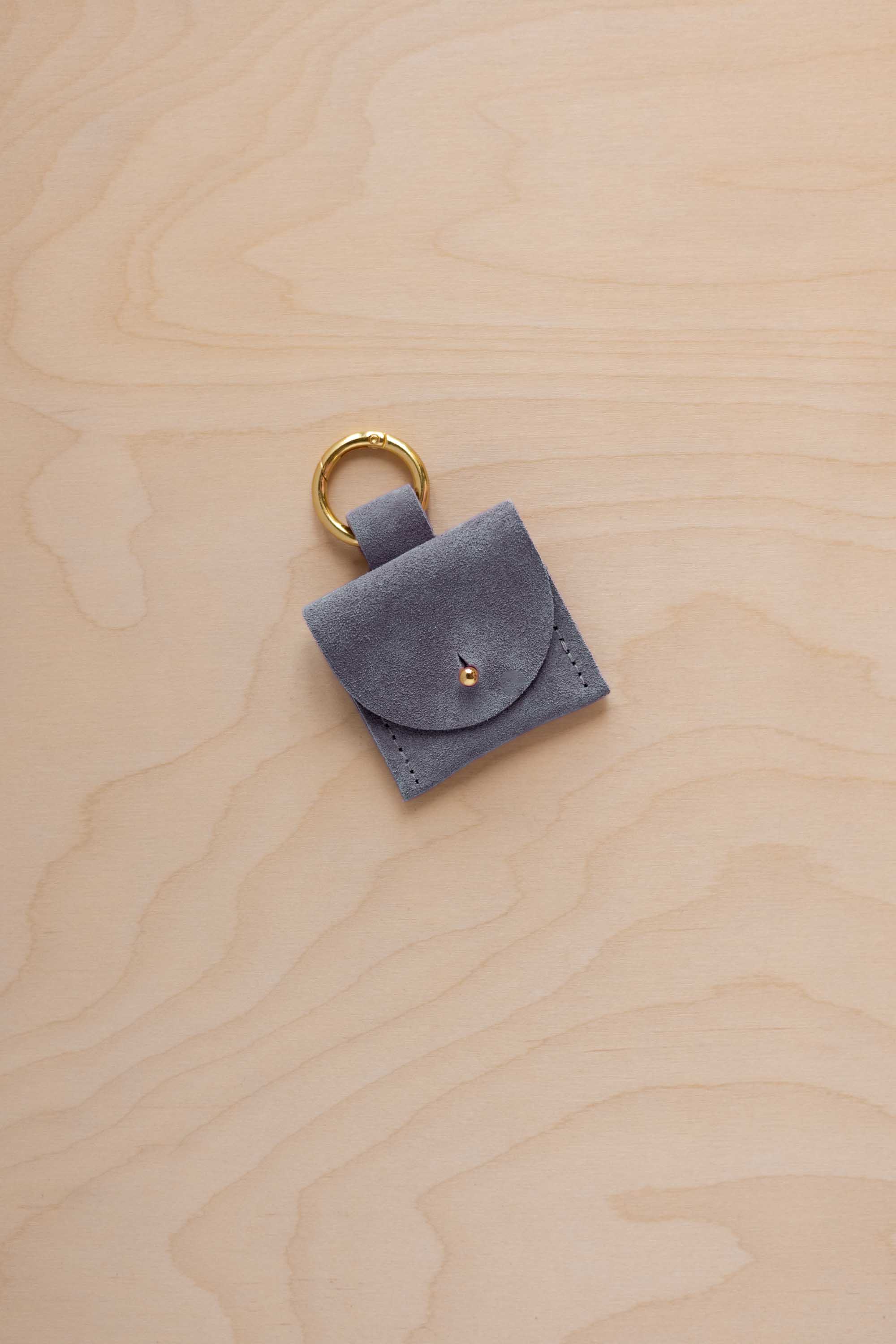 A small dusky blue coloured suede pouch bag charm with a gold coloured clip ring fastening, displayed on a light wood grain background.