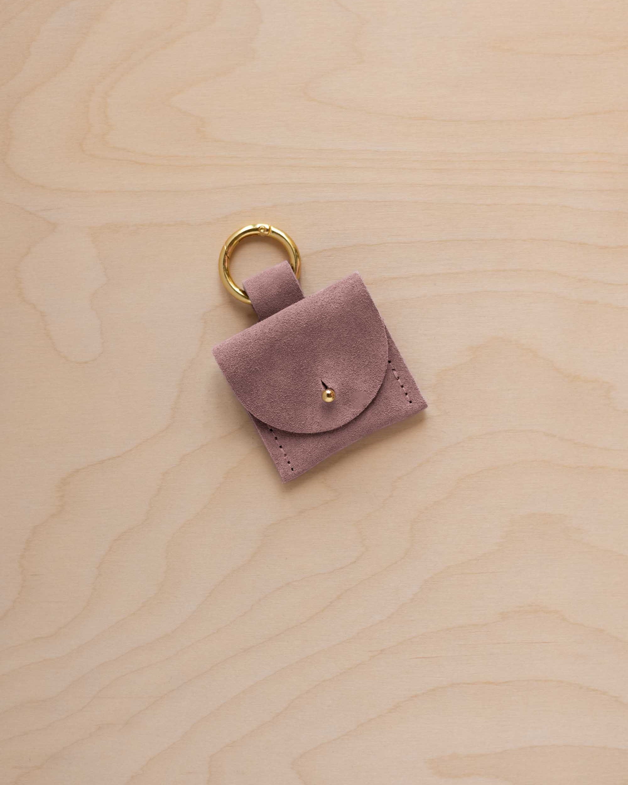 A small dusty mauve suede pouch bag charm with a gold coloured clip ring fastening, displayed on a light wood grain background.
