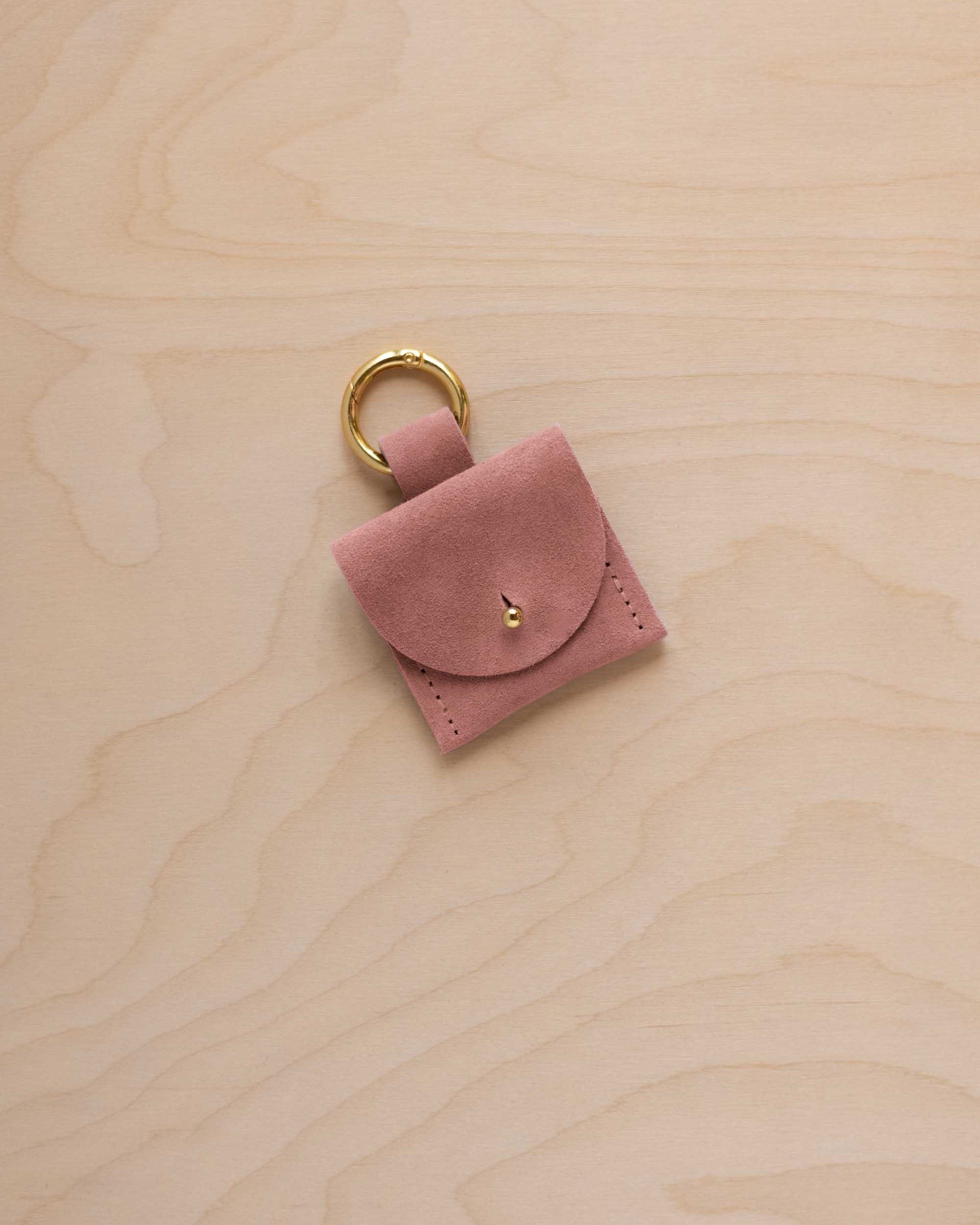 A small dusty rose coloured pale pink suede pouch bag charm with a gold coloured clip ring fastening, displayed on a light wood grain background.