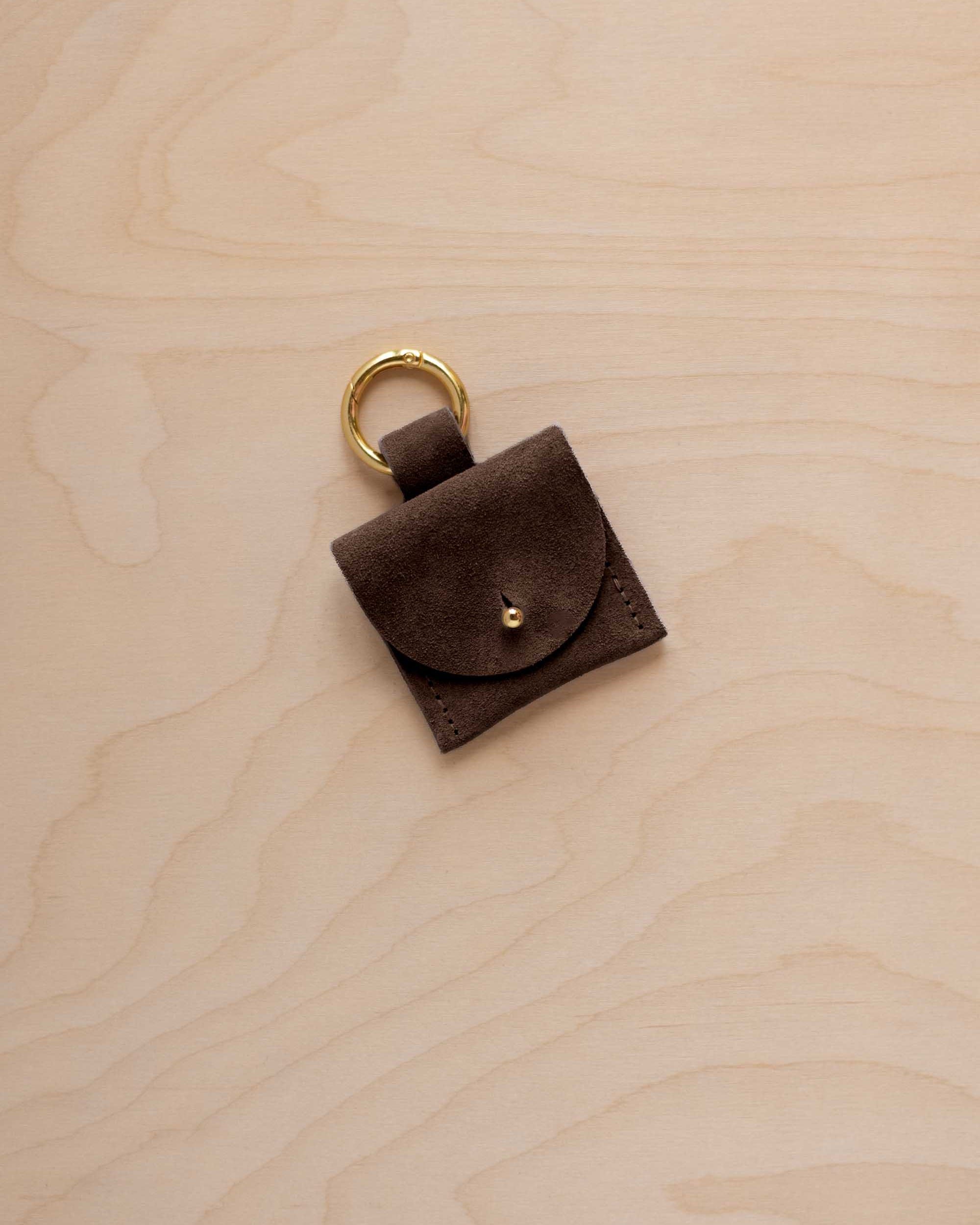 A small espresso brown coloured suede pouch bag charm with a gold coloured clip ring fastening, displayed on a light wood grain background.