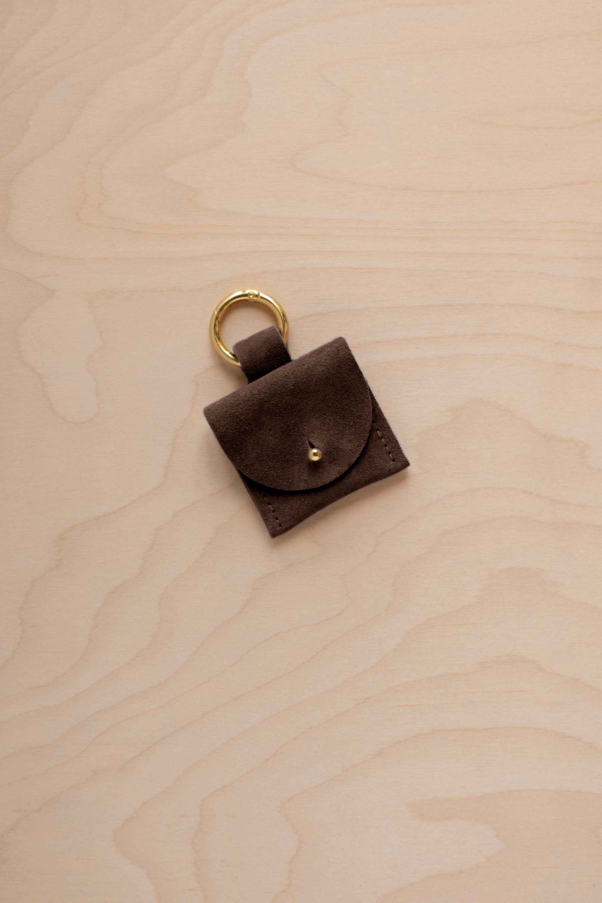 A small espresso brown coloured suede pouch bag charm with a gold coloured clip ring fastening, displayed on a light wood grain background.