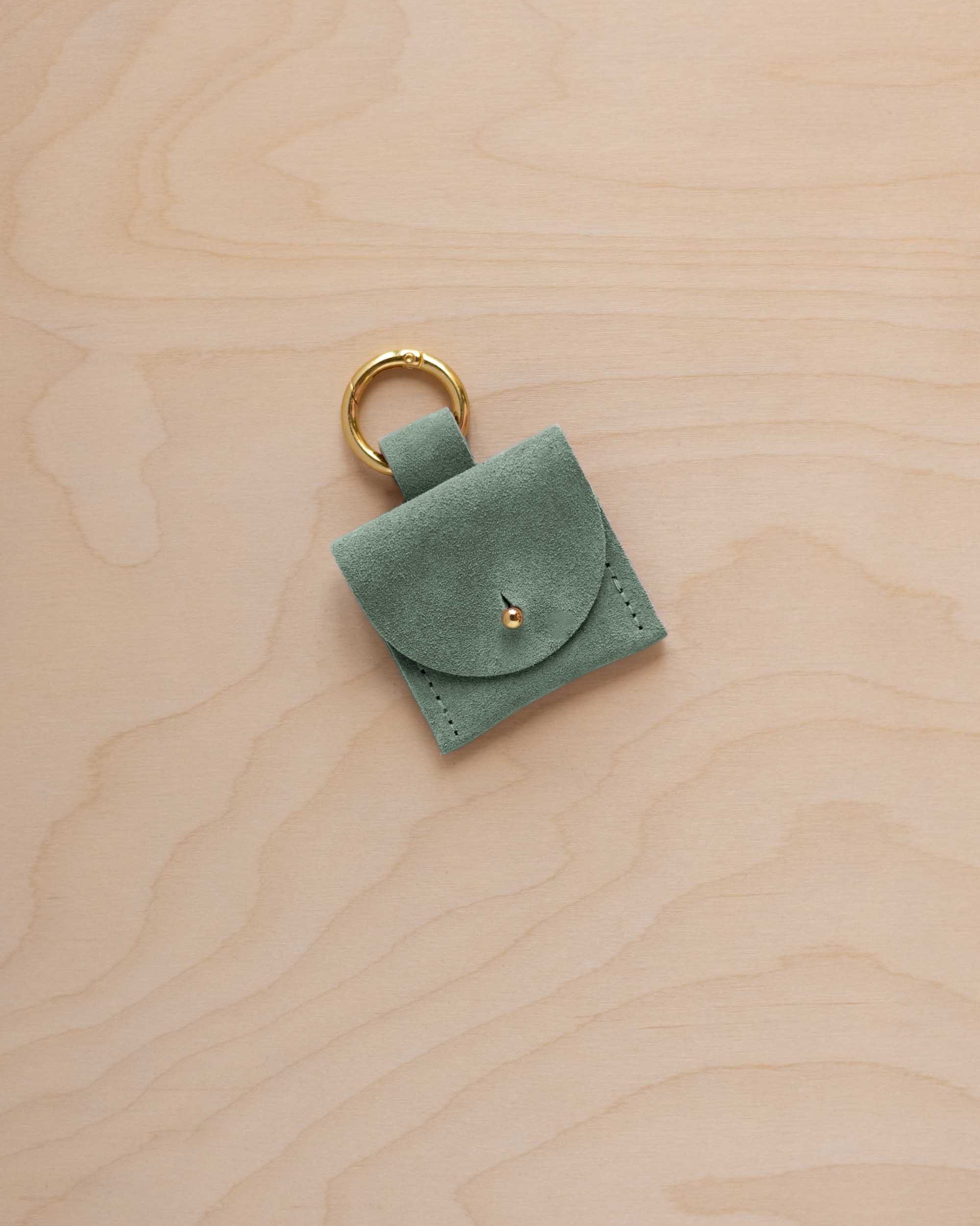 A small eucalyptus coloured sage green suede pouch bag charm with a gold coloured clip ring fastening, displayed on a light wood grain background.