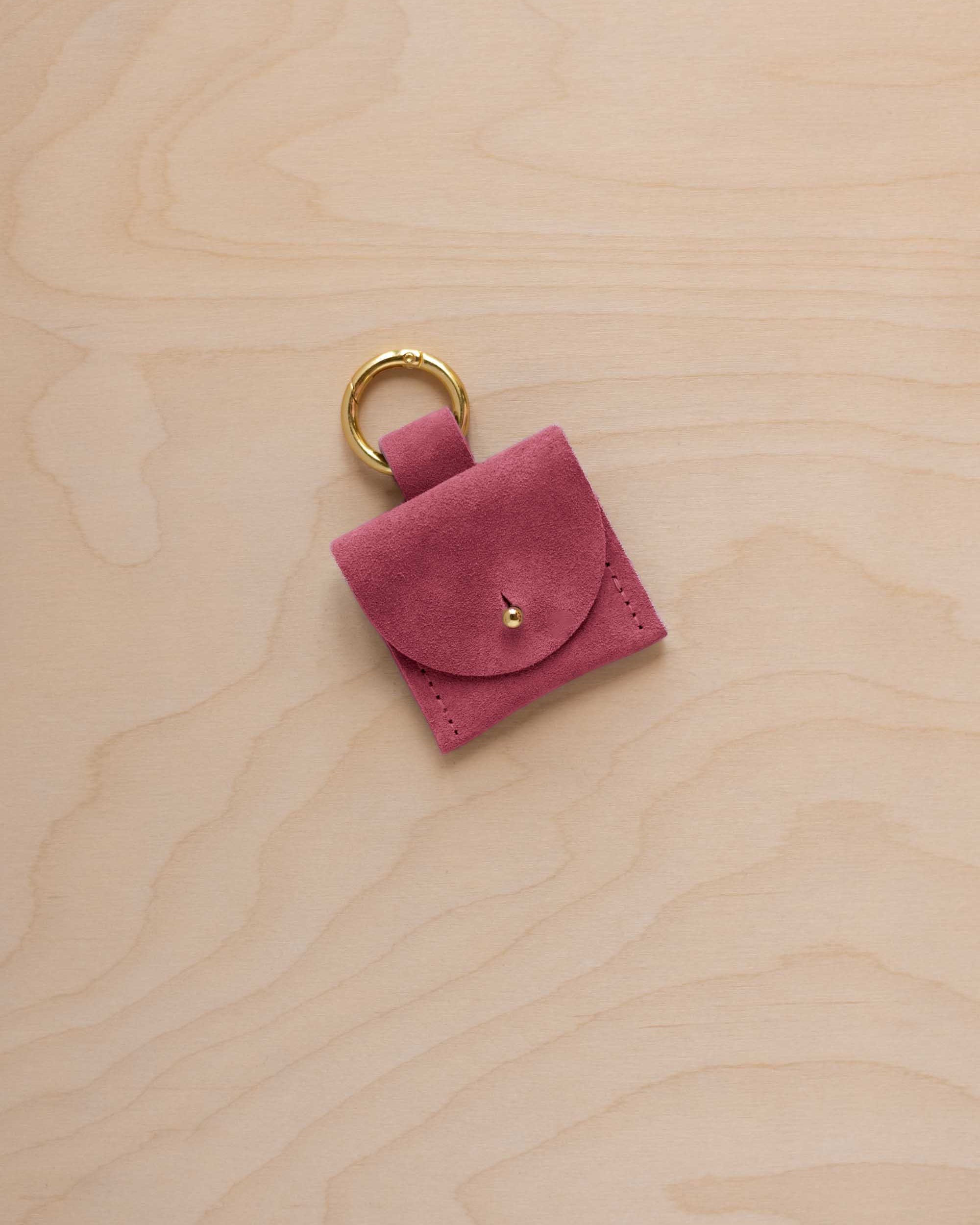A small faded rose pink coloured suede pouch bag charm with a gold coloured clip ring fastening, displayed on a light wood grain background.