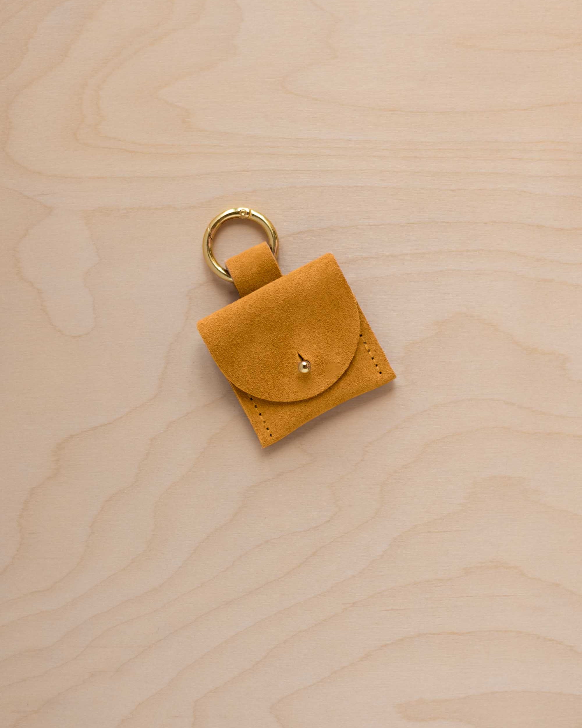 A small golden ochre mustard yellow yellow suede pouch bag charm with a gold coloured clip ring fastening, displayed on a light wood grain background.