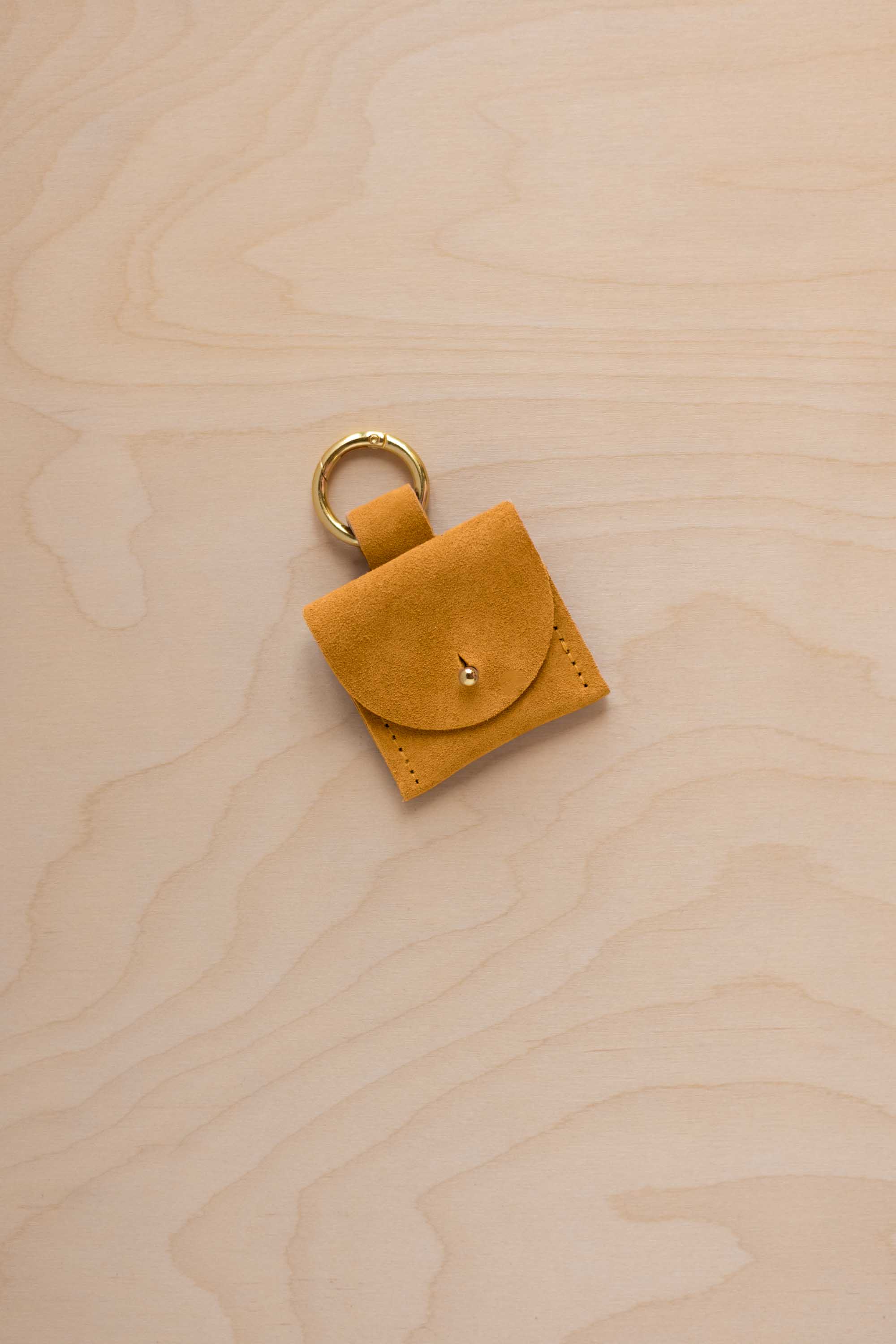 A small golden ochre mustard yellow yellow suede pouch bag charm with a gold coloured clip ring fastening, displayed on a light wood grain background.