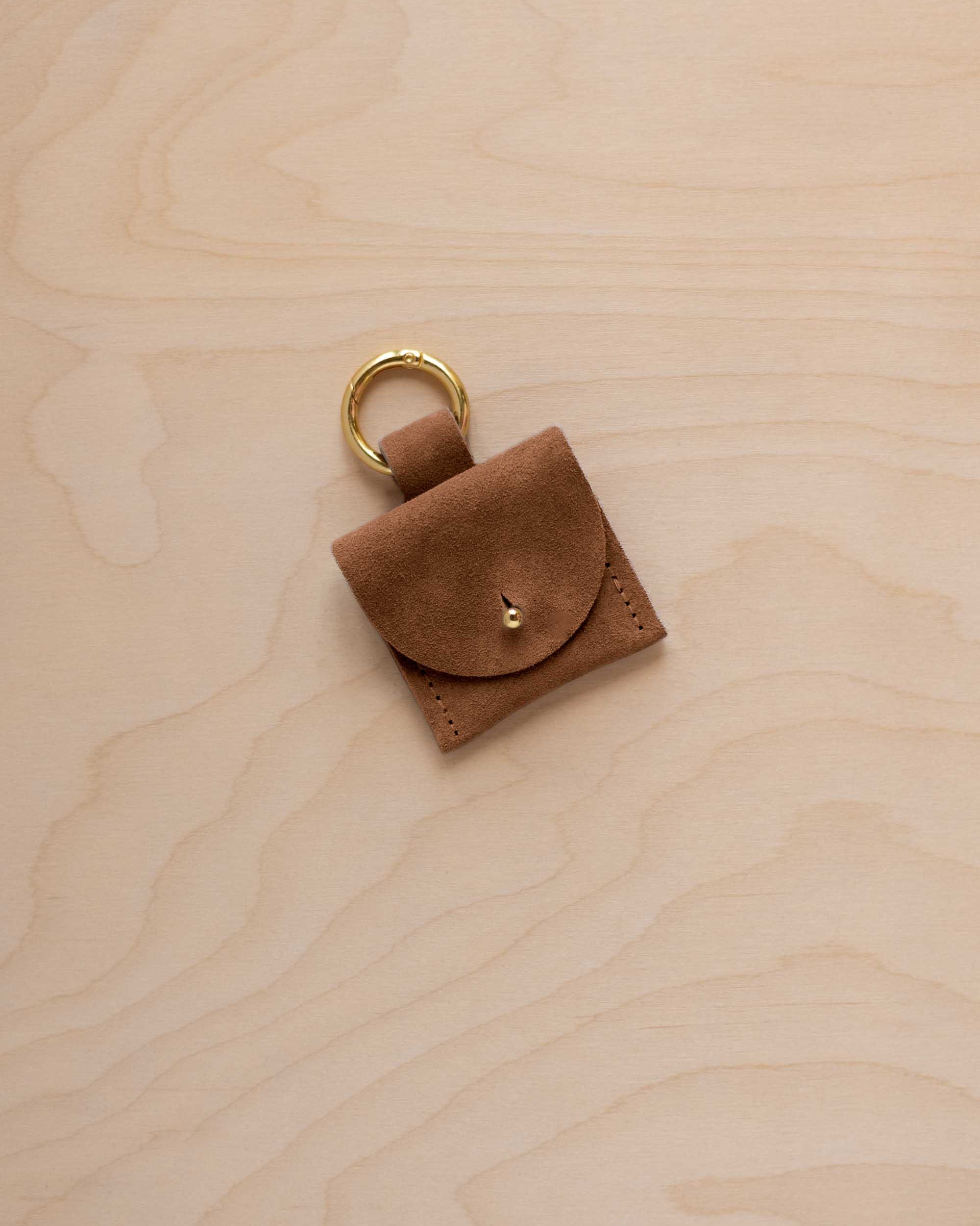 A small mocha brown coloured suede pouch bag charm with a gold coloured clip ring fastening, displayed on a light wood grain background.