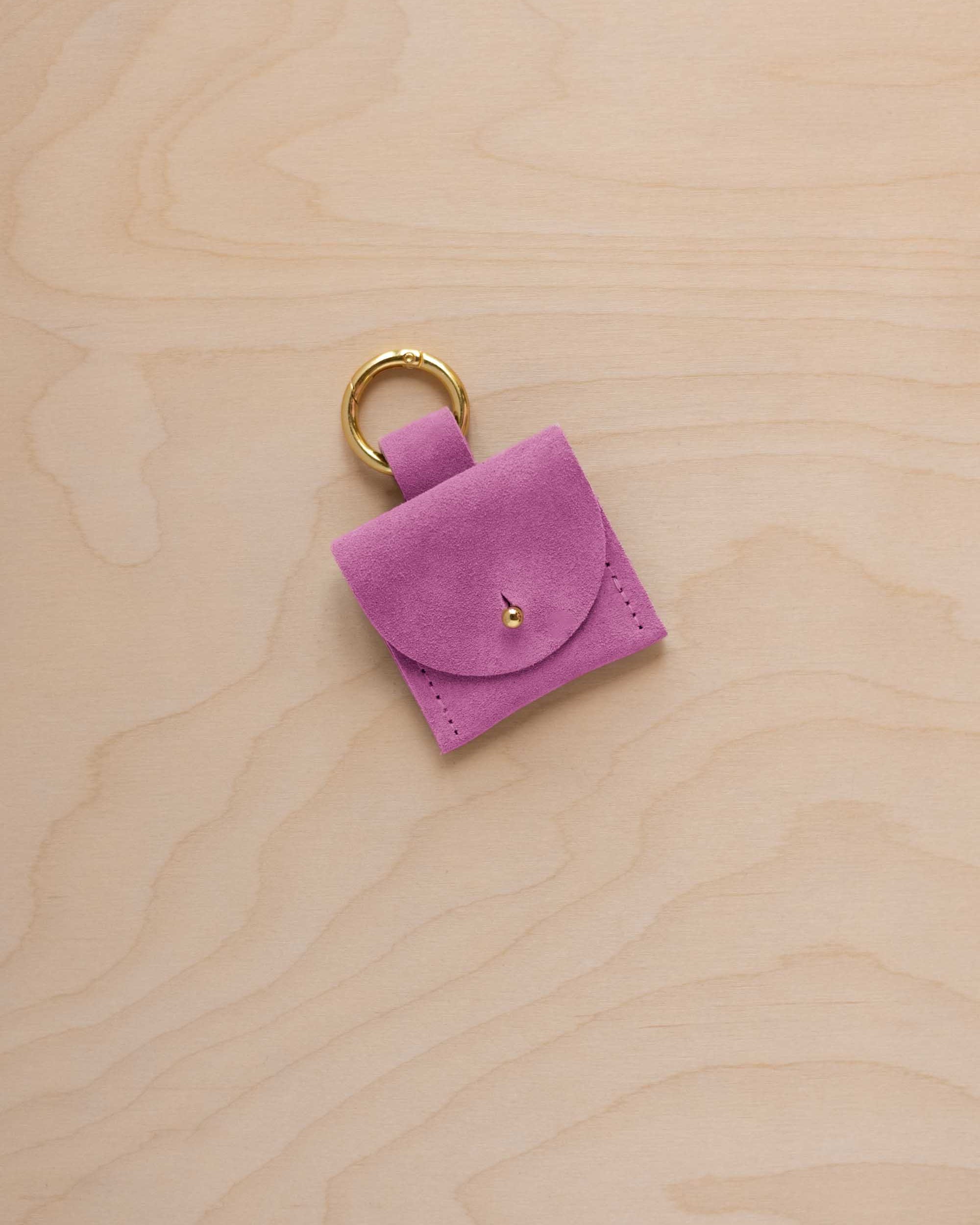 A small peony pink suede pouch bag charm with a gold coloured clip ring fastening, displayed on a light wood grain background.
