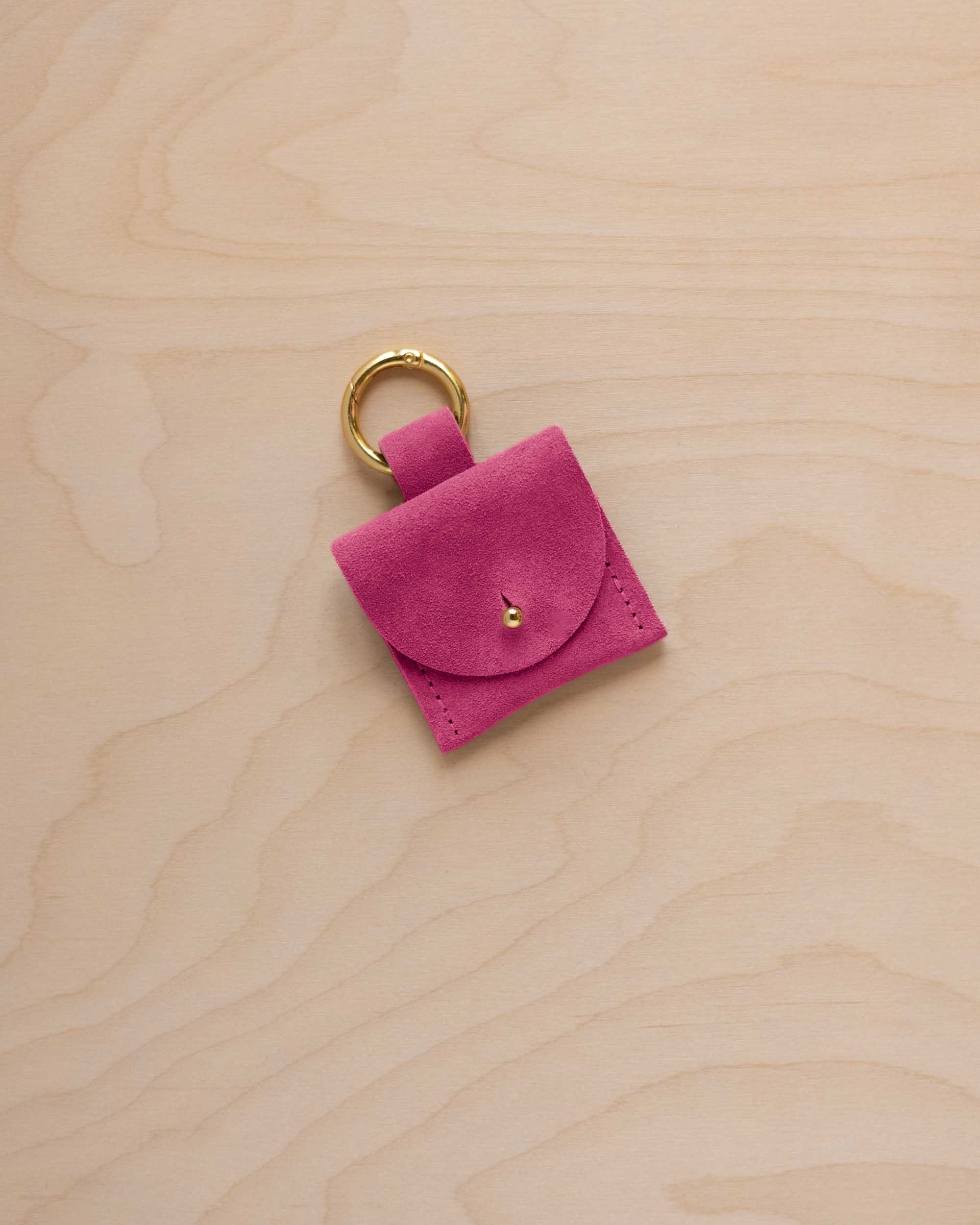 A small raspberry pink coloured suede pouch bag charm with a gold coloured clip ring fastening, displayed on a light wood grain background.
