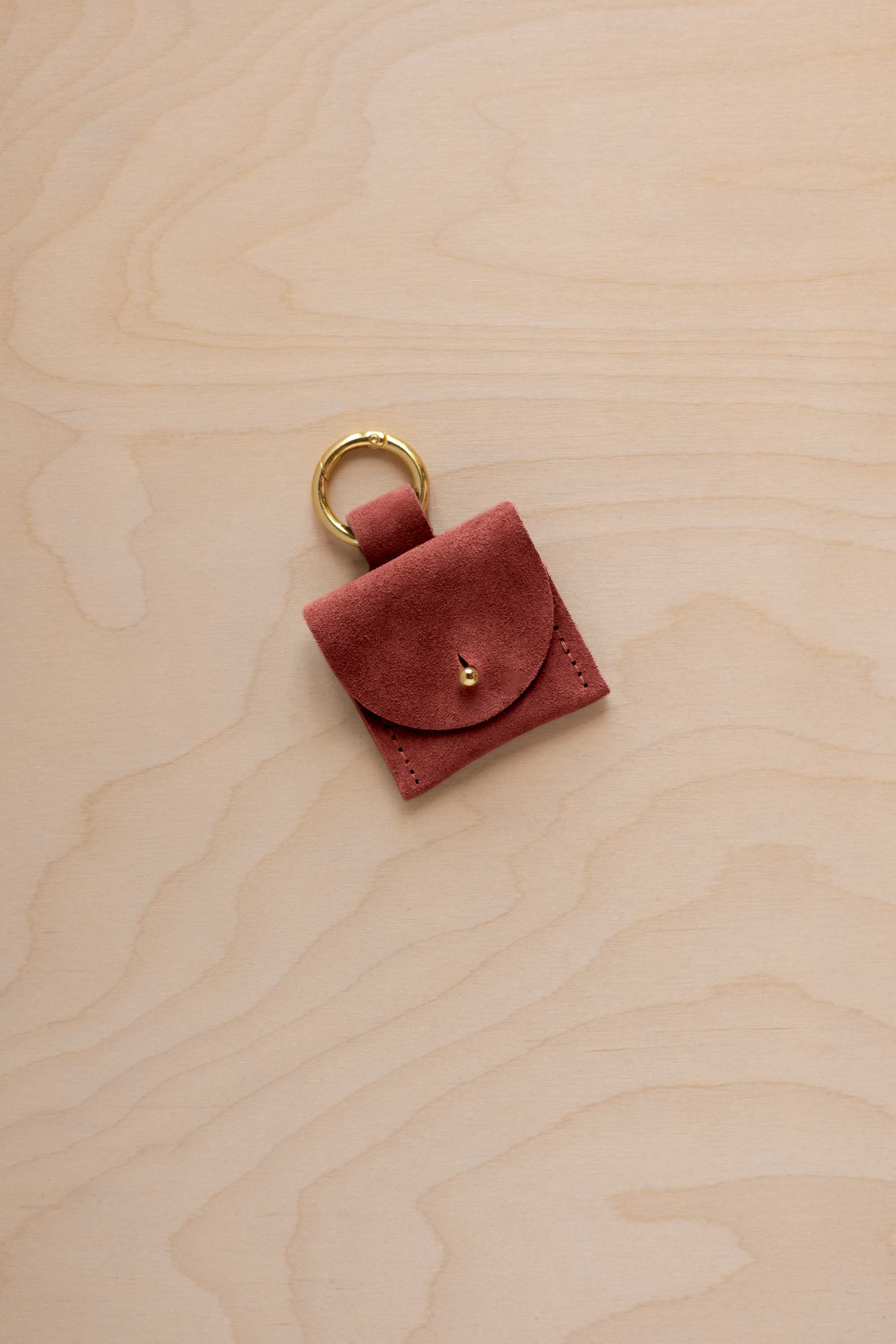 A small rust red coloured suede pouch bag charm with a gold coloured clip ring fastening, displayed on a light wood grain background.