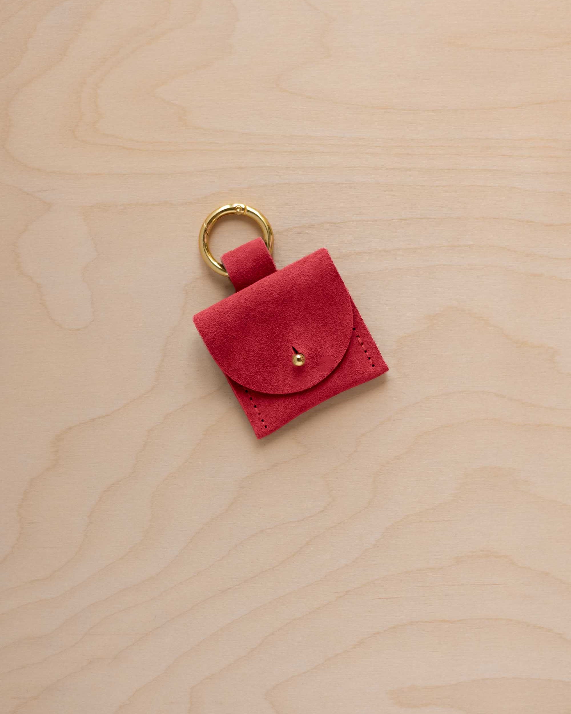 A small scarlet red coloured suede pouch bag charm with a gold coloured clip ring fastening, displayed on a light wood grain background.