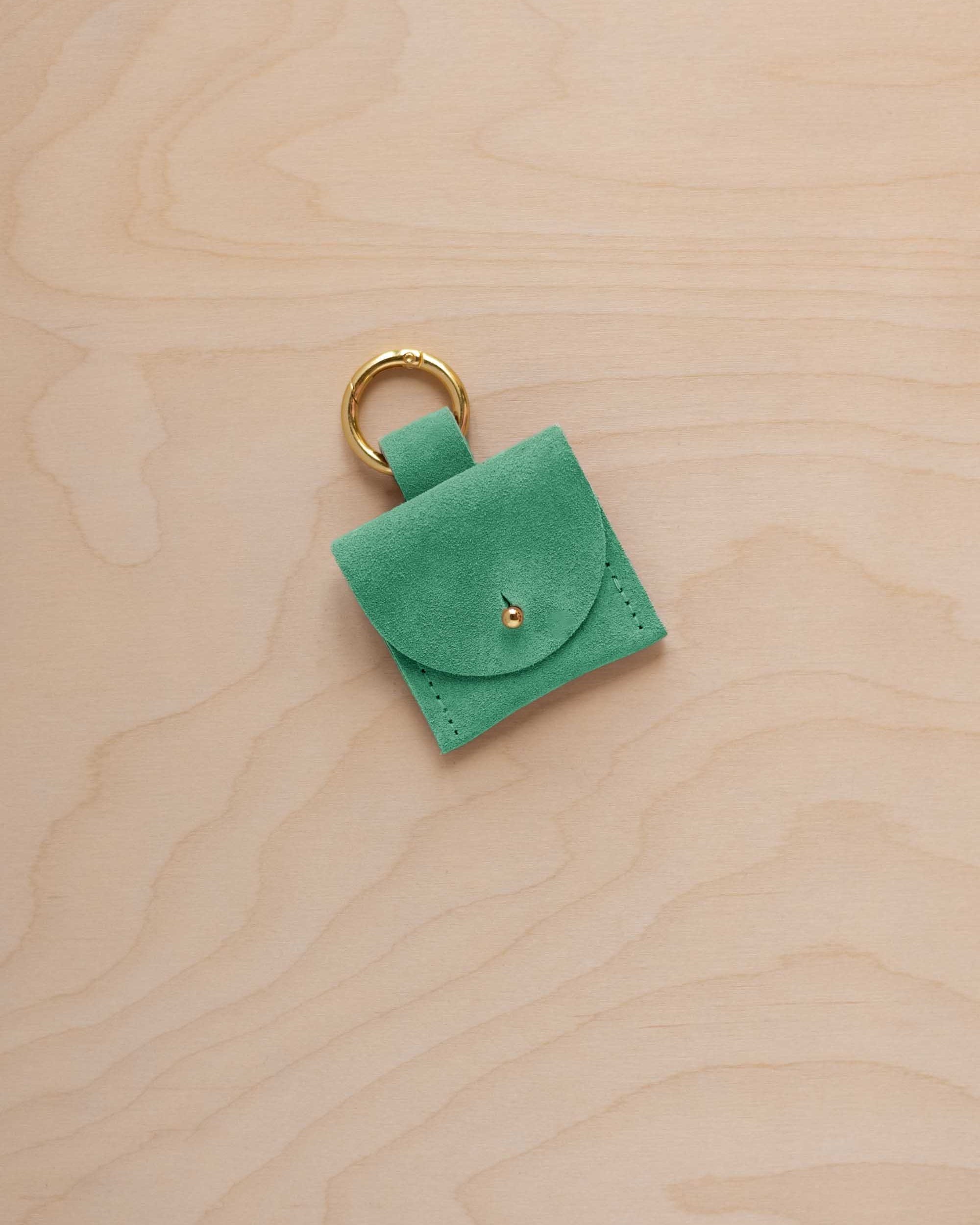 A small seafoam green turquoise coloured suede pouch bag charm with a gold coloured clip ring fastening, displayed on a light wood grain background.