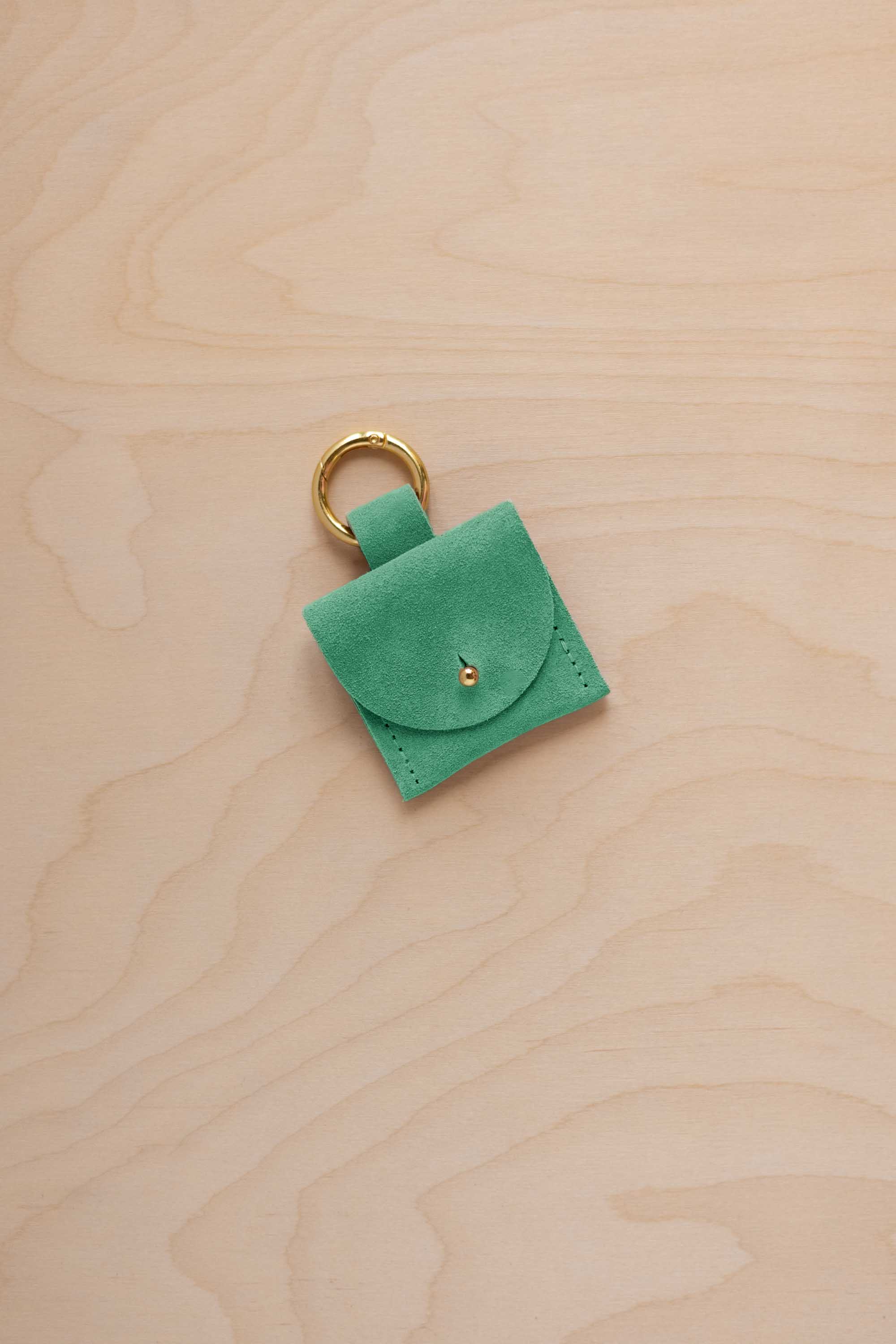 A small seafoam green turquoise coloured suede pouch bag charm with a gold coloured clip ring fastening, displayed on a light wood grain background.