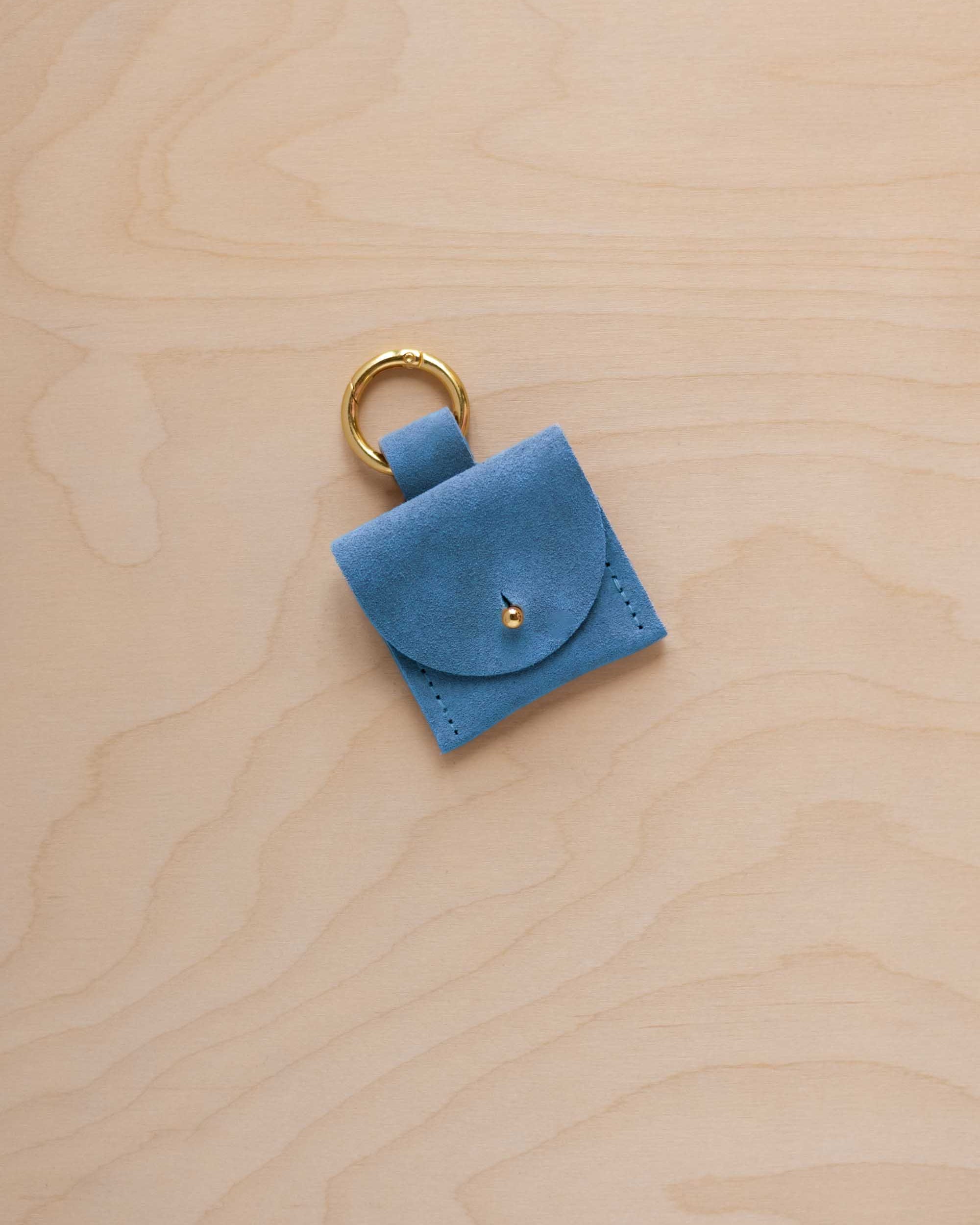 A small sky blue coloured suede pouch bag charm with a gold coloured clip ring fastening, displayed on a light wood grain background.