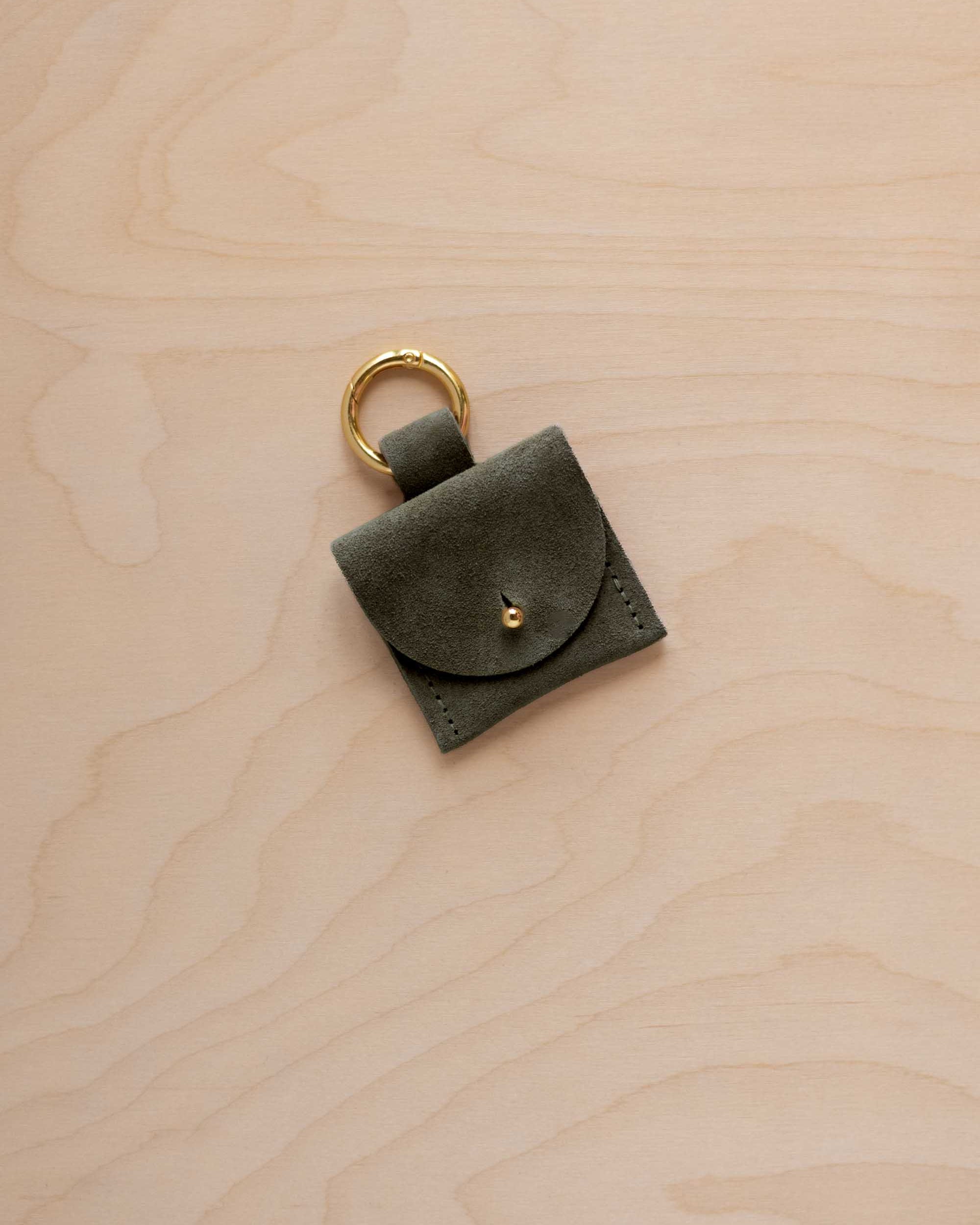 A small soft olive green coloured suede pouch bag charm with a gold coloured clip ring fastening, displayed on a light wood grain background.