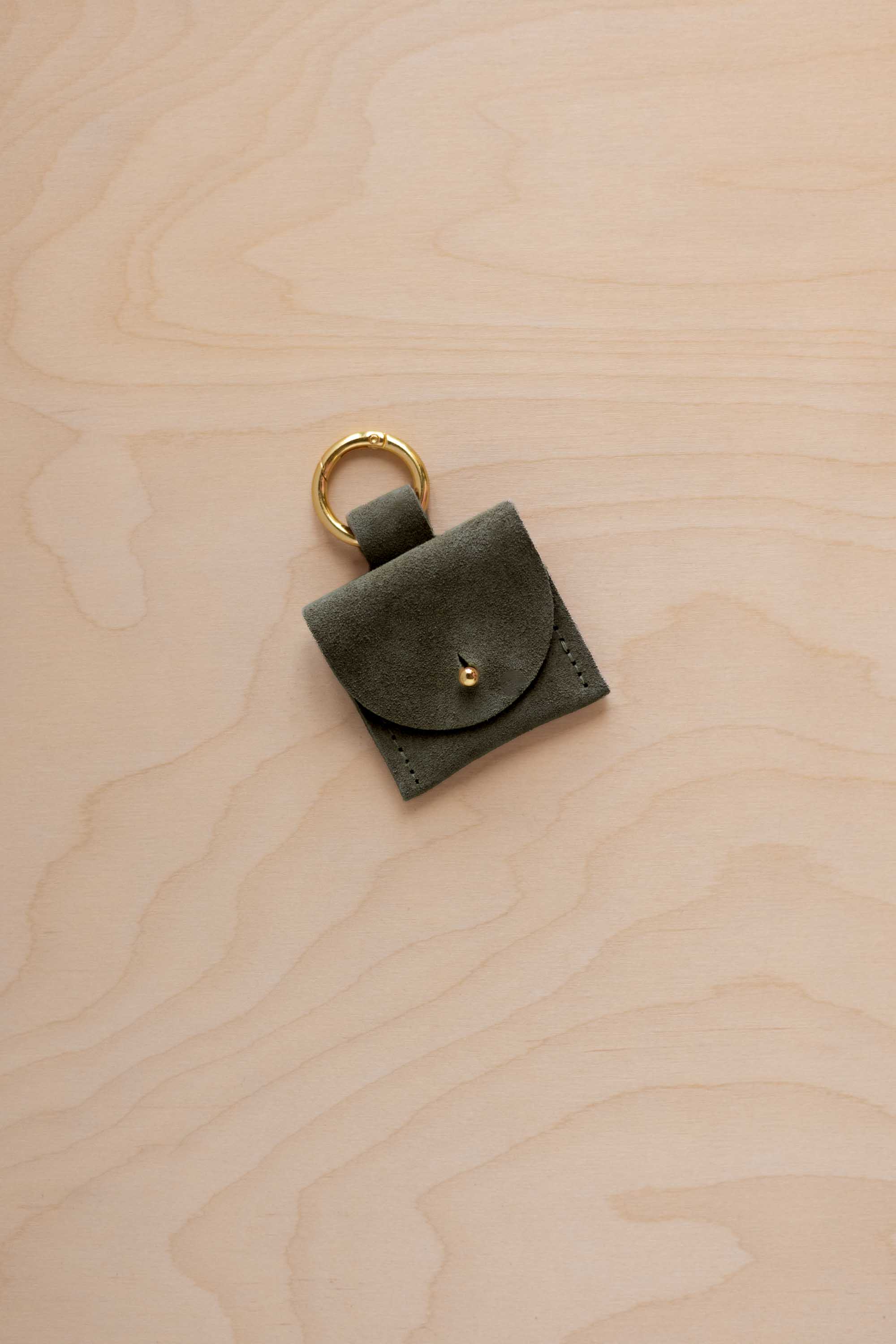 A small soft olive green coloured suede pouch bag charm with a gold coloured clip ring fastening, displayed on a light wood grain background.
