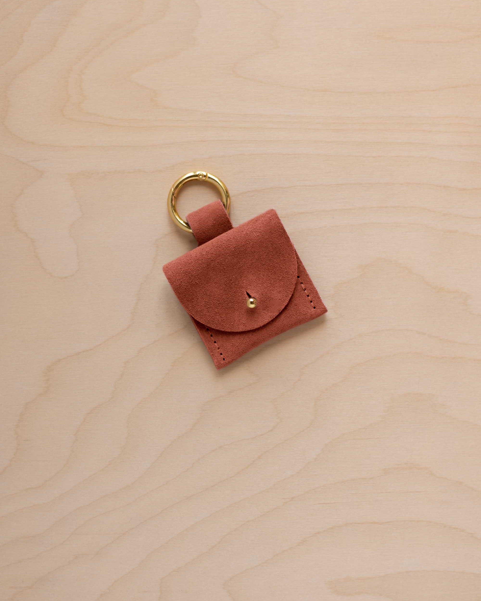 A small terracotta brown coloured suede pouch bag charm with a gold coloured clip ring fastening, displayed on a light wood grain background.
