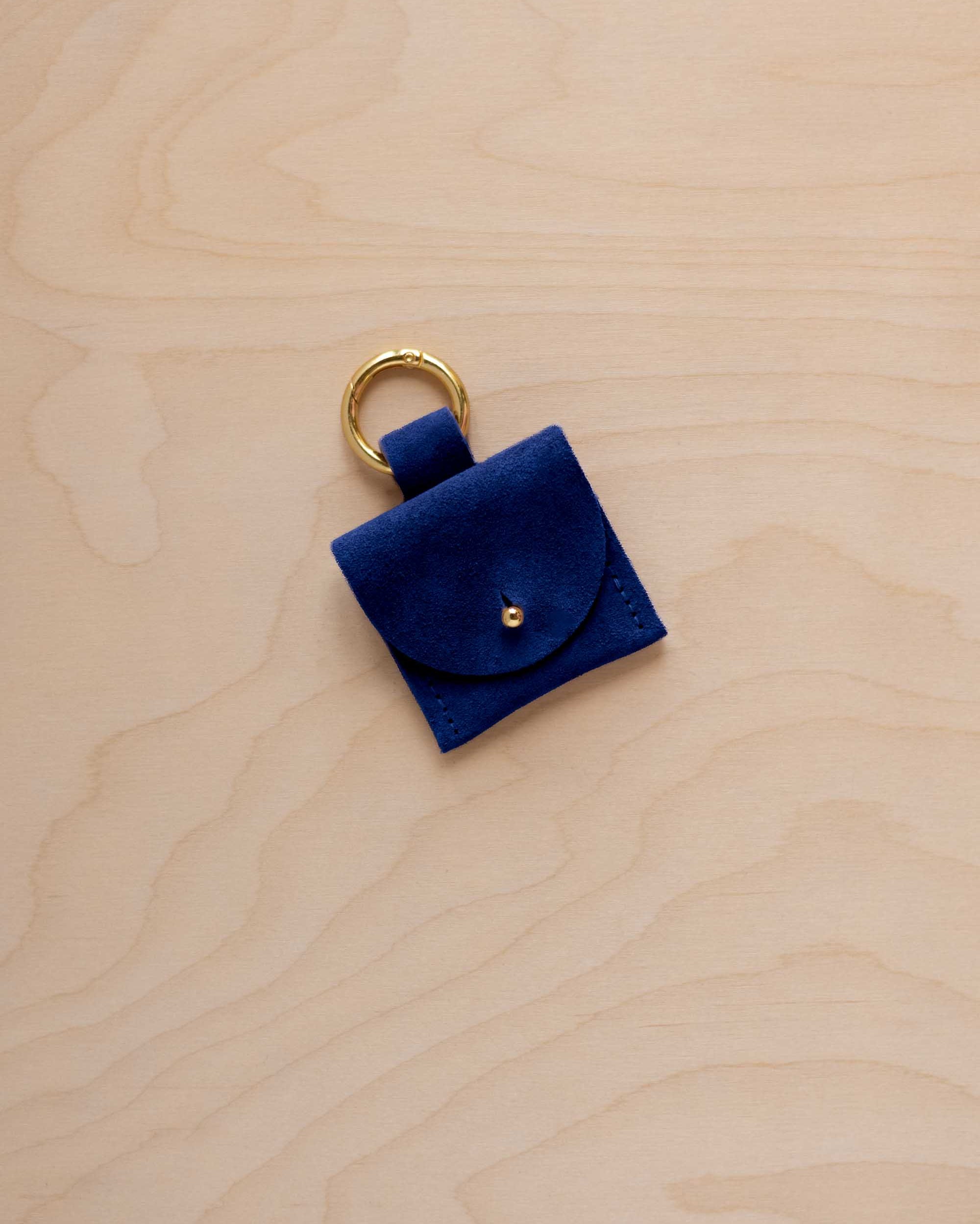 A small ultramarine blue coloured suede pouch bag charm with a gold coloured clip ring fastening, displayed on a light wood grain background.