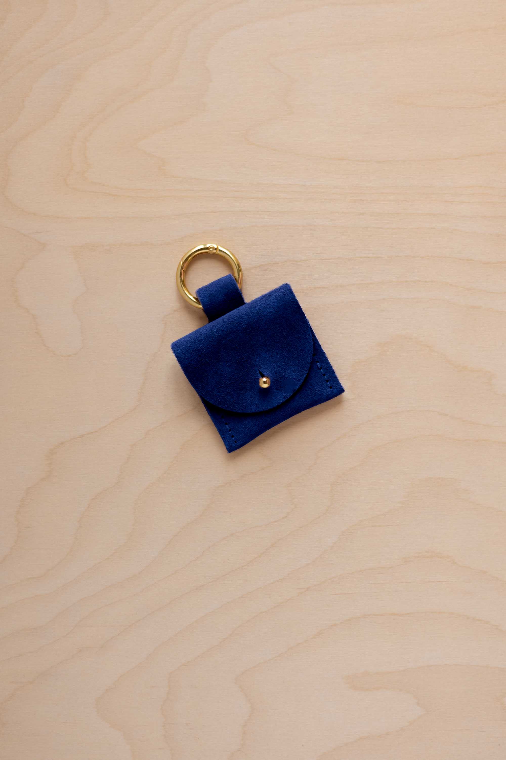 A small ultramarine blue coloured suede pouch bag charm with a gold coloured clip ring fastening, displayed on a light wood grain background.